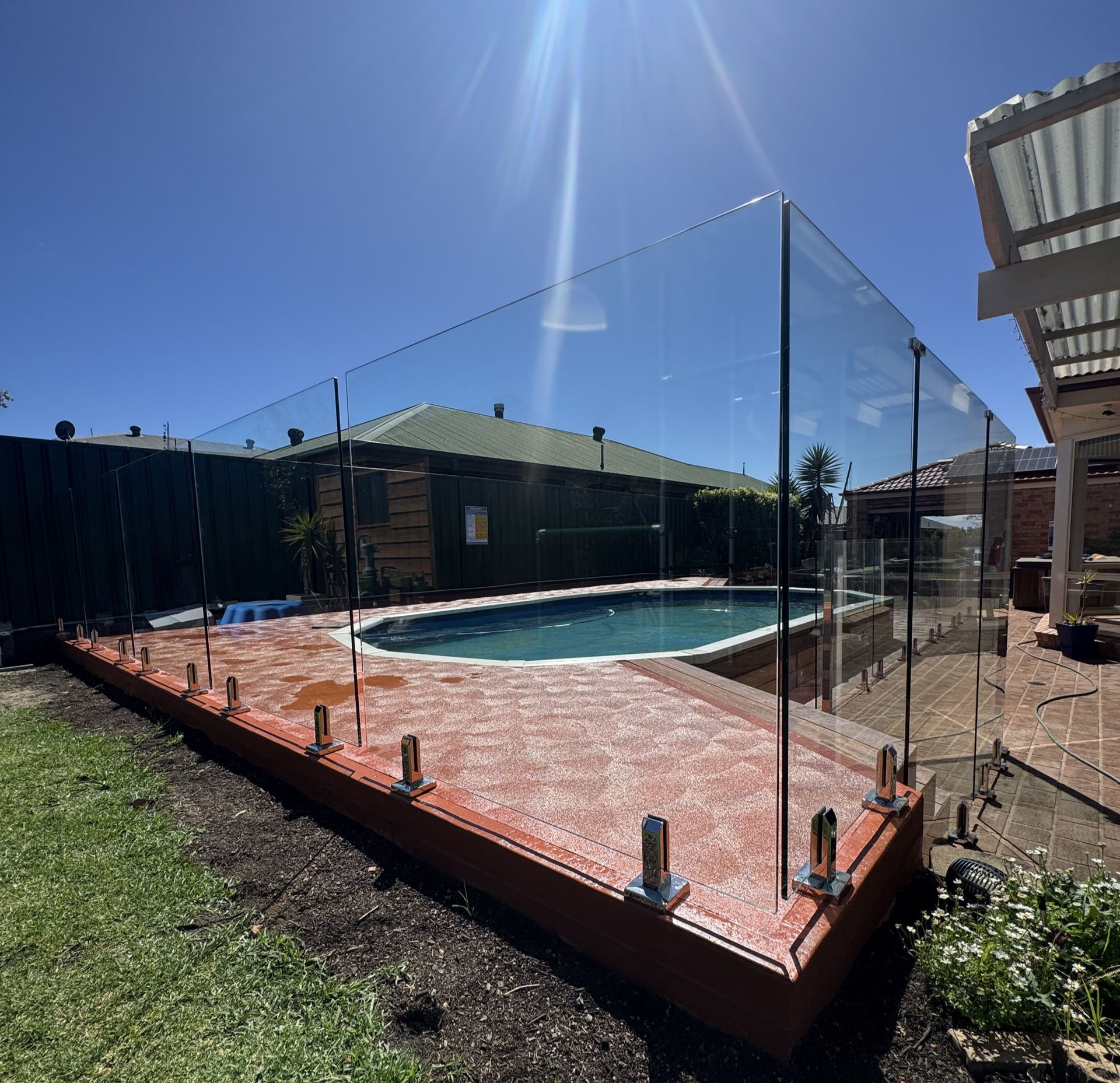 Pool area with glass fencing, wooden deck, and blue sky. — CAW Services in Badagarang, NSW