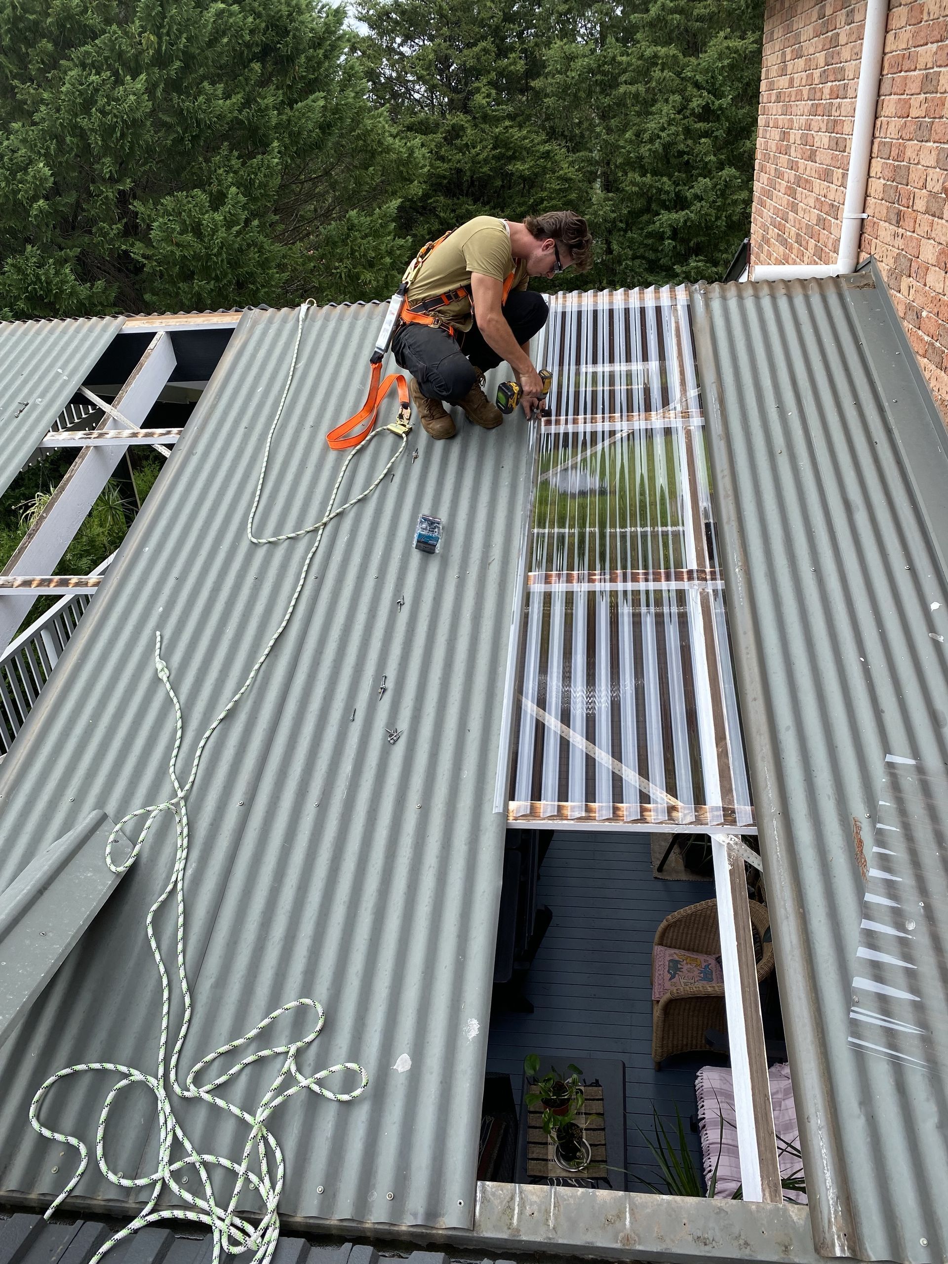 Person working on a corrugated metal roof, installing translucent panels. Safety harness and rope visible  — CAW Services in Badagarang, NSW