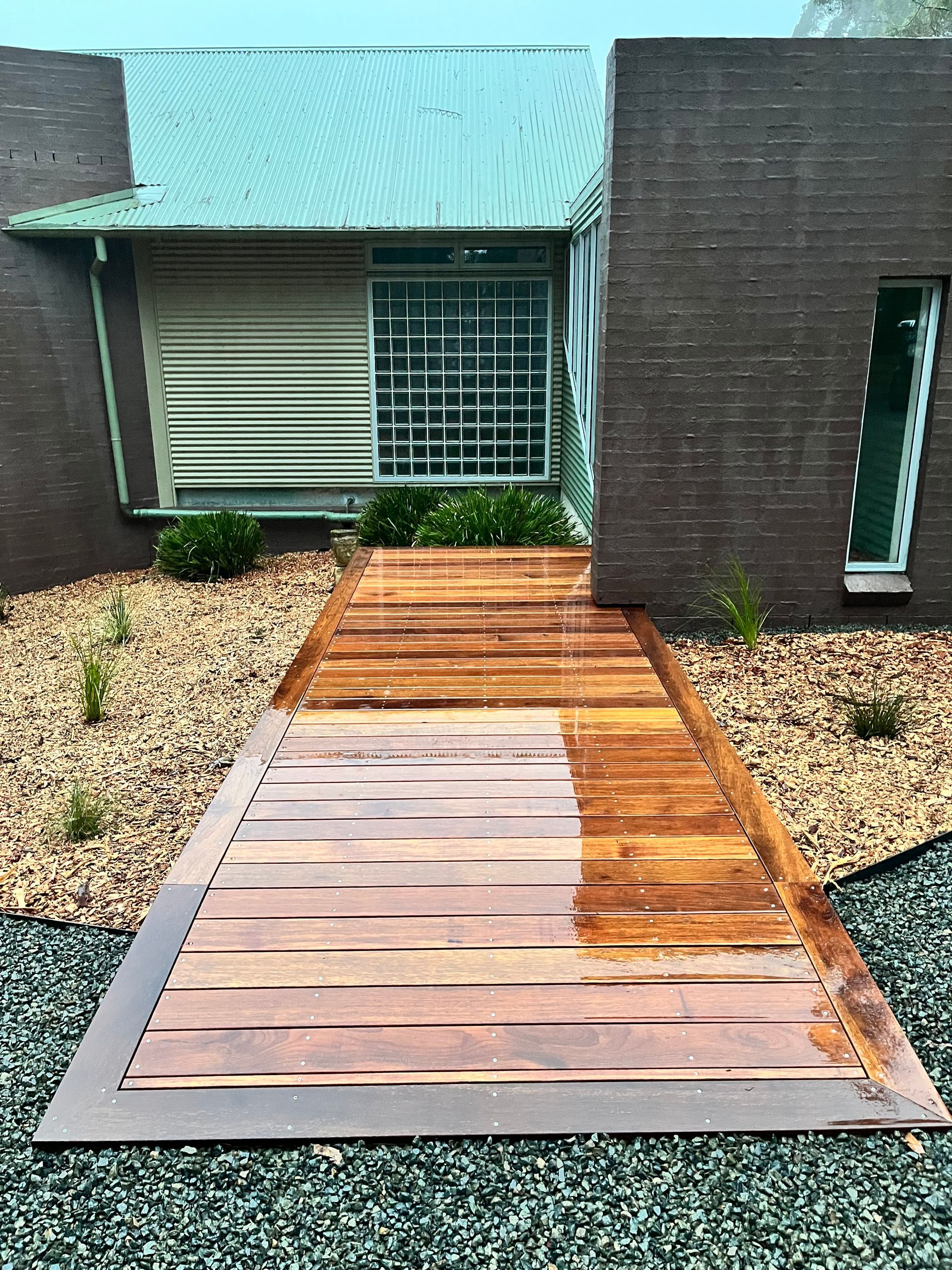 Wooden walkway leading to a modern building with a green roof, bordered by gravel and small plants. — CAW Services in Badagarang, NSW