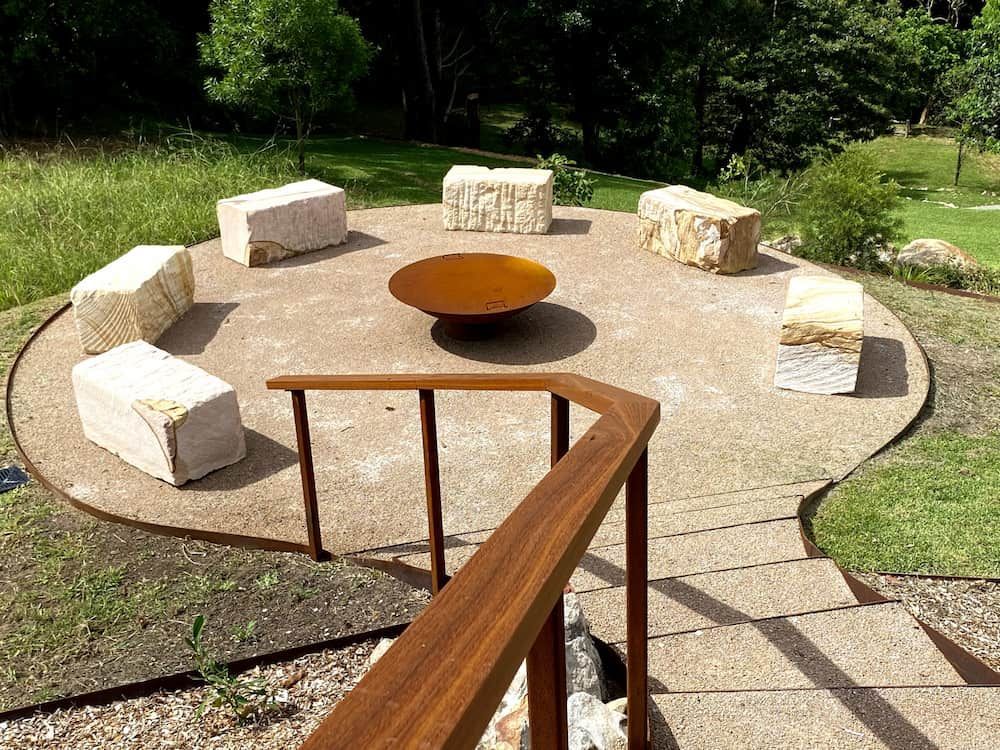 Stone Seating Encircles a Rusty Fire Pit on a Gravel Patio, With a Wooden Railing — CAW Services in Badagarang, NSW