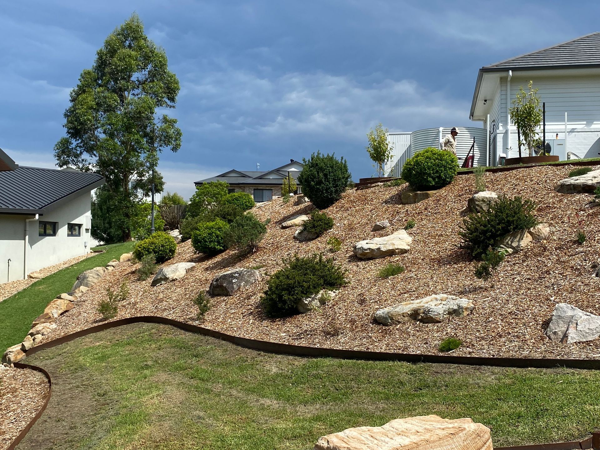 Landscaped hillside with mulch, shrubs, and rocks, under a cloudy sky, near houses  — CAW Services in Badagarang, NSW