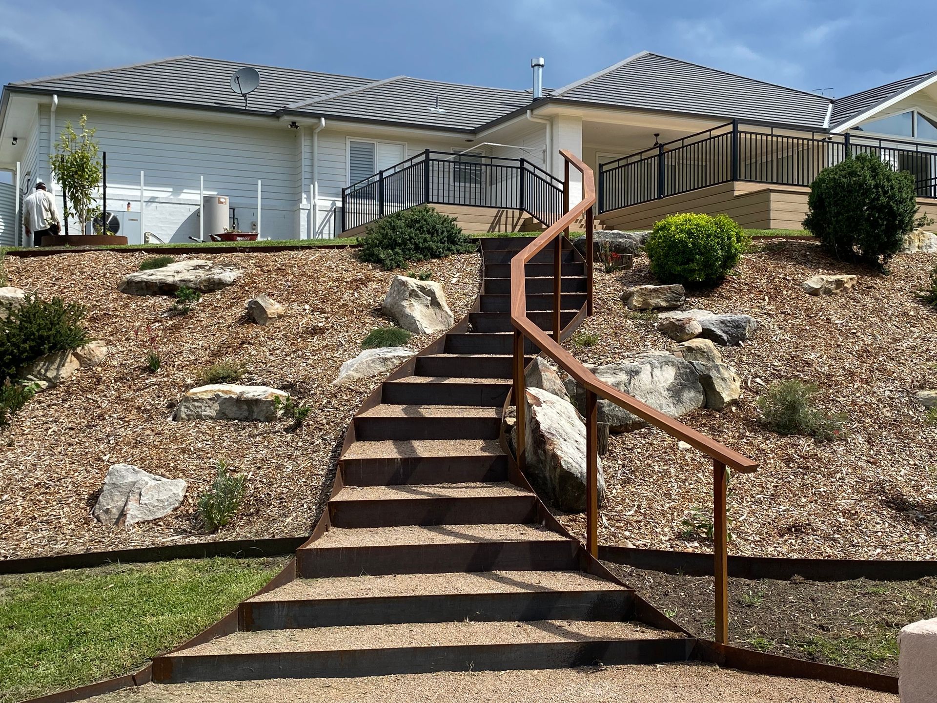 Stone steps with brown railing lead up a hillside to a house with striped awnings. — CAW Services in Badagarang, NSW