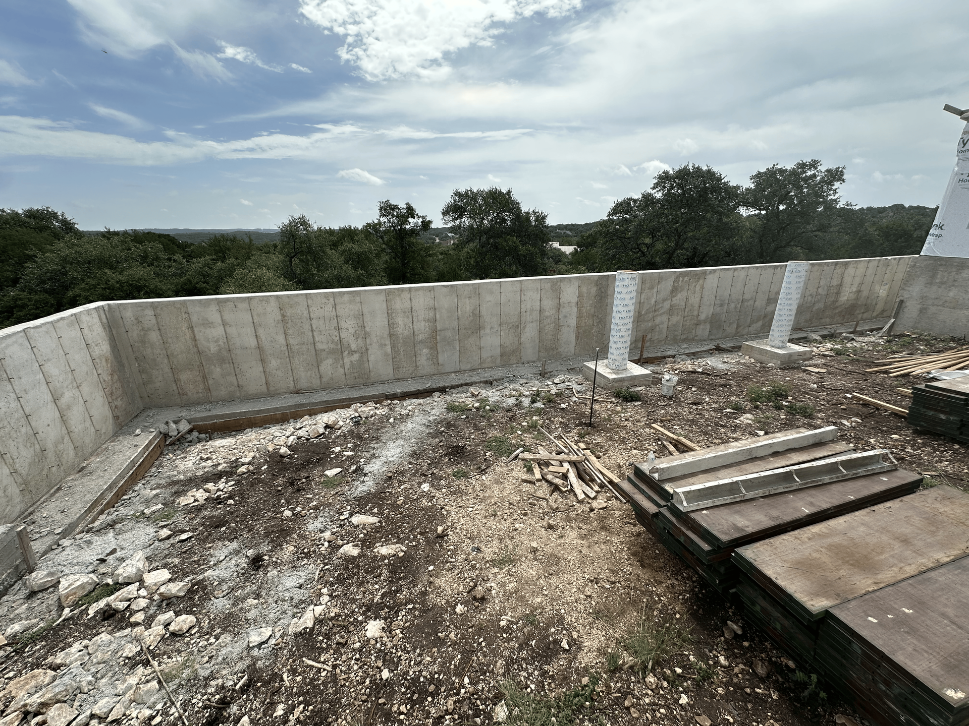 Construction site: concrete wall with exposed ground and surrounding trees under a cloudy sky.