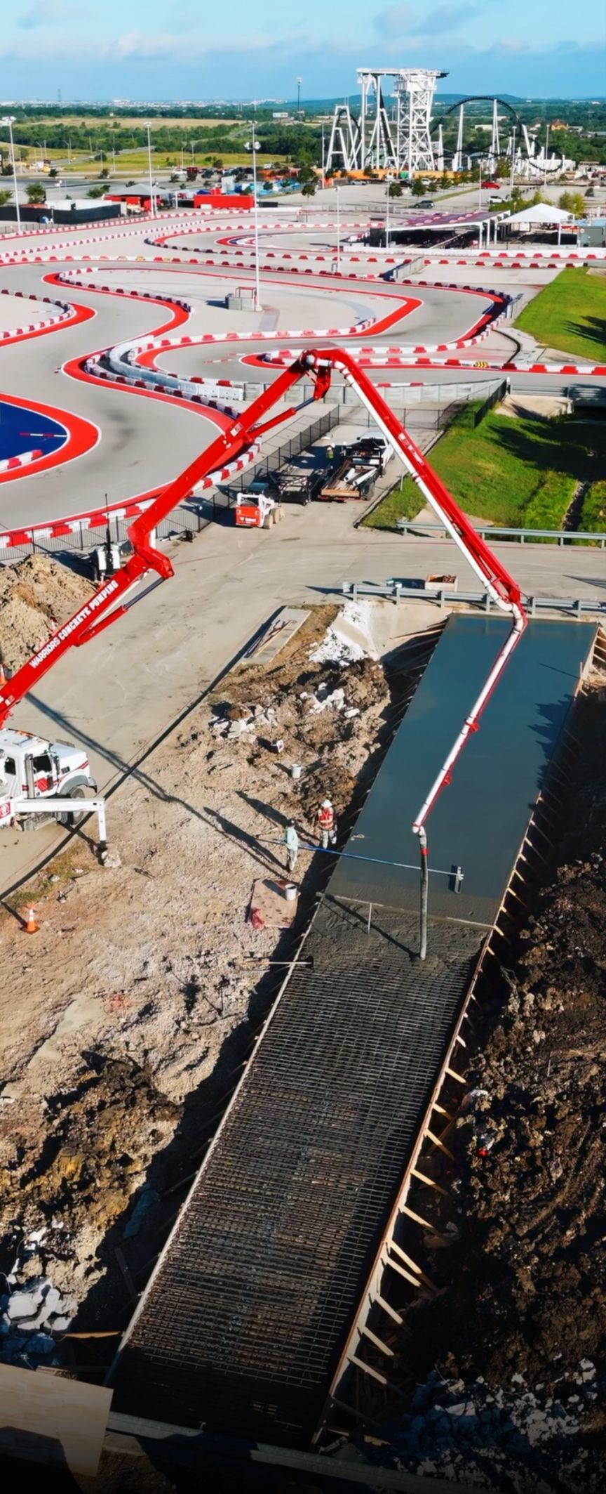 Construction of a racetrack with concrete being poured by a pump truck. A track and industrial facilities are in the background.