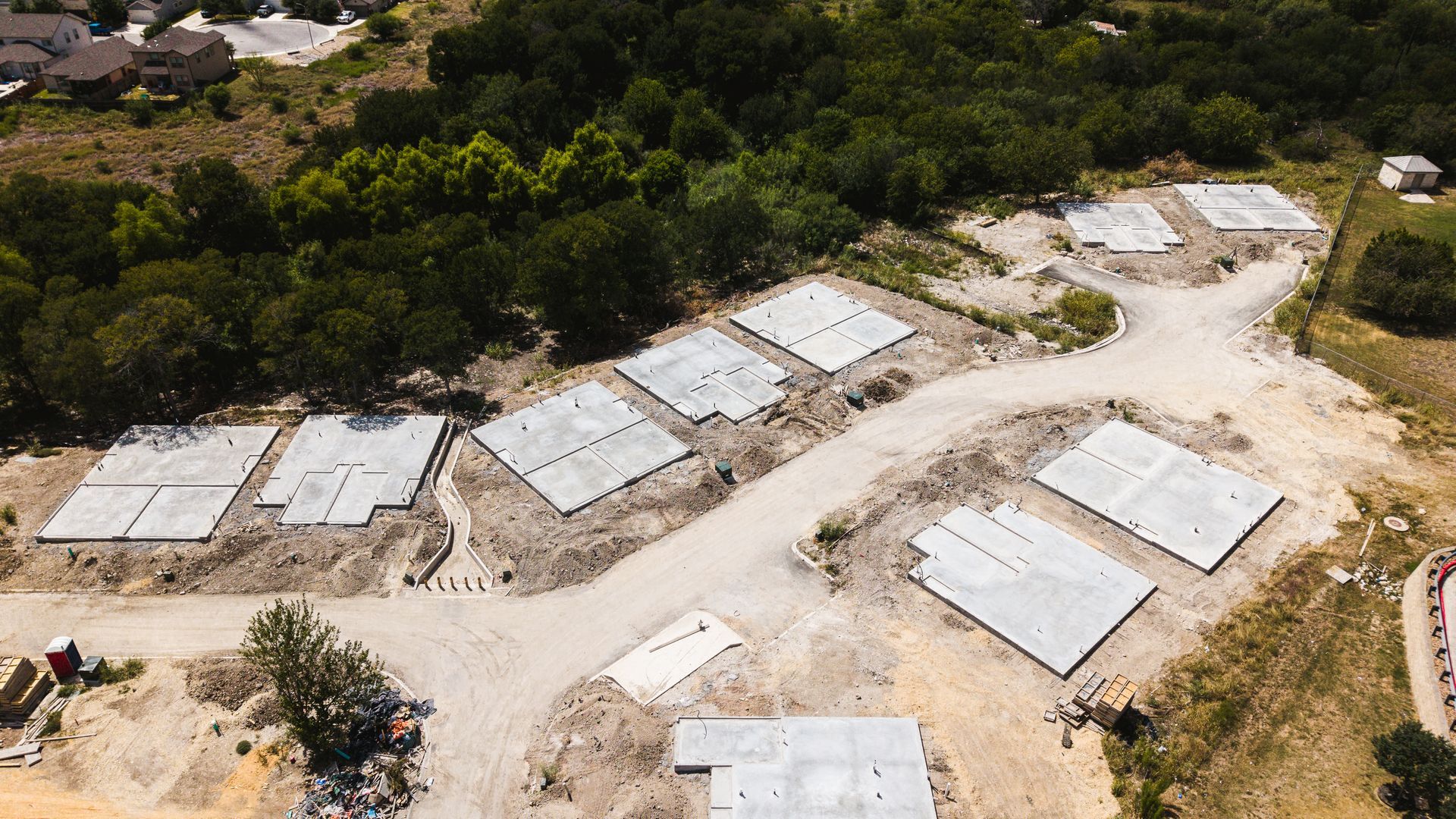 Aerial view of concrete foundations laid out for houses in a construction site, surrounded by dirt and trees.