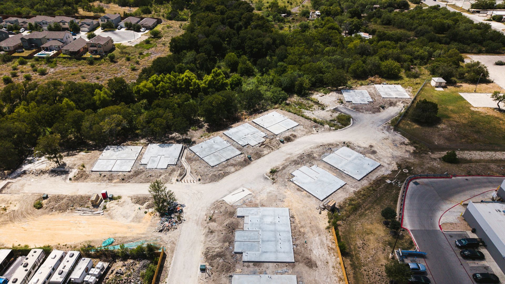 Aerial view of a construction site with concrete foundations and dirt roads, surrounded by trees and buildings.