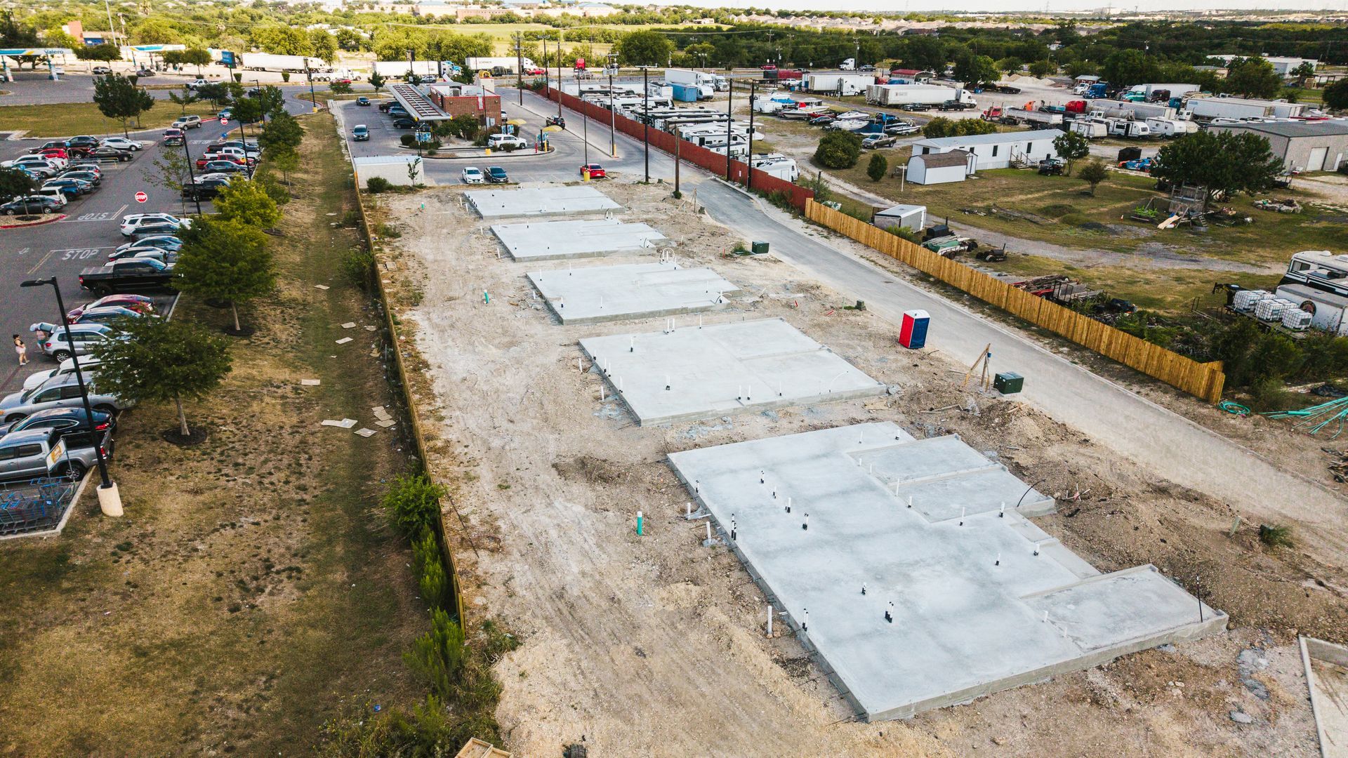 Construction site with concrete foundations, surrounded by dirt and a fence. Commercial buildings in the background.
