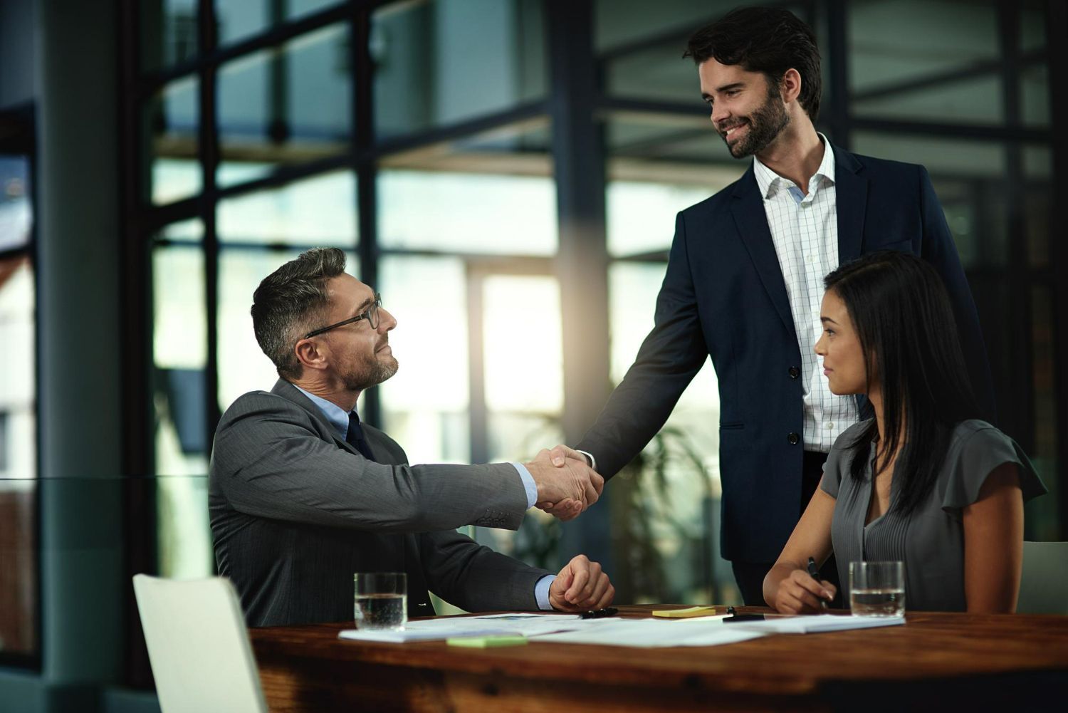 Two men in suits shaking hands at a table, with a woman in business attire looking on.