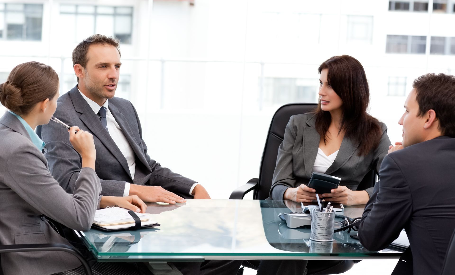 Four business professionals in suits seated around a conference table, engaged in a discussion. Bright, modern office setting.