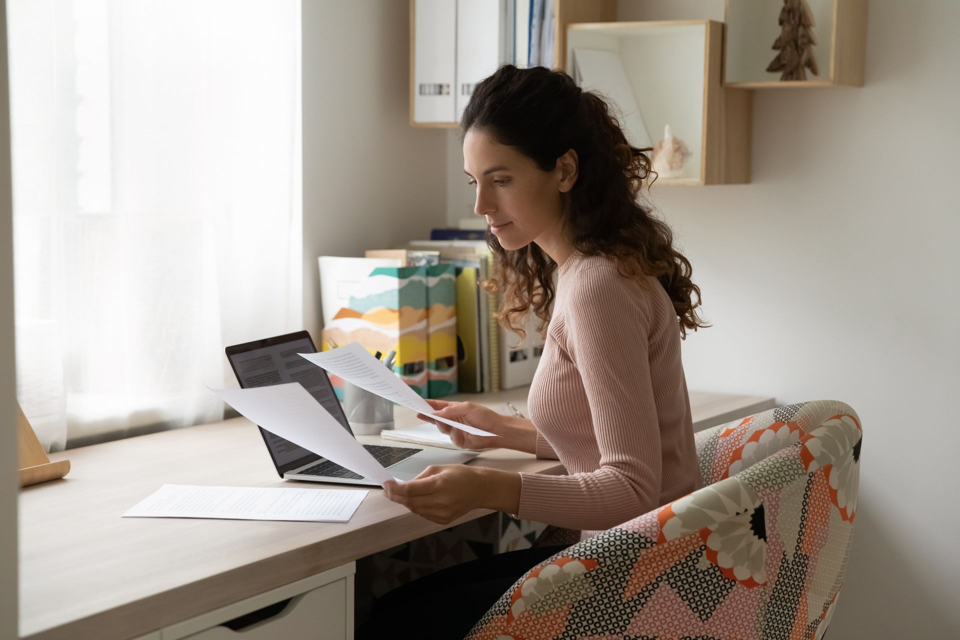 Woman in pink sweater working at a desk, reviewing papers, with a laptop and window in the background.