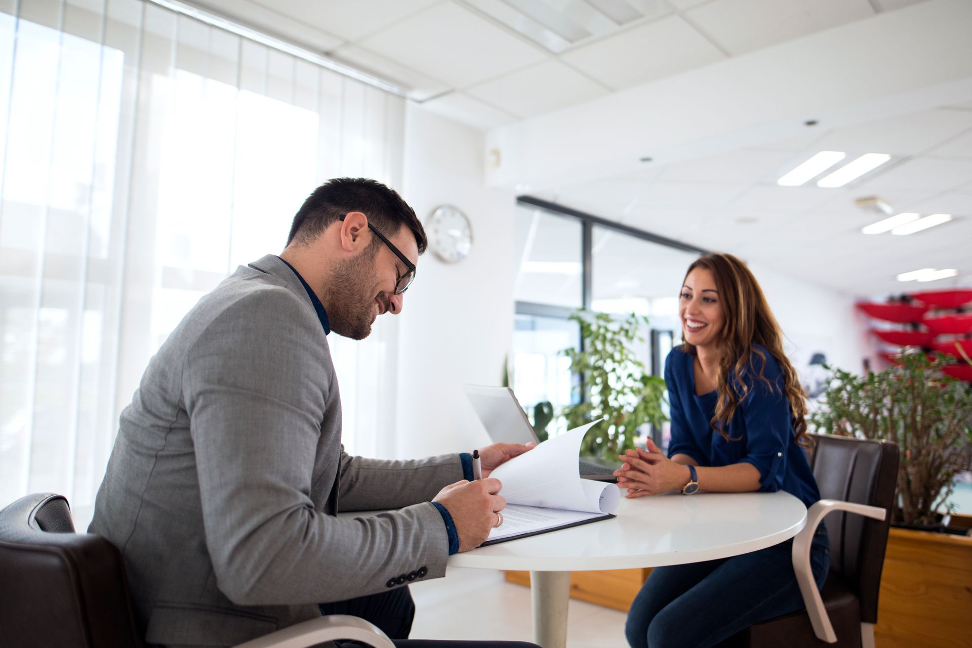 A man in a gray blazer reviews documents with a smiling woman at a round table in a well-lit office.
