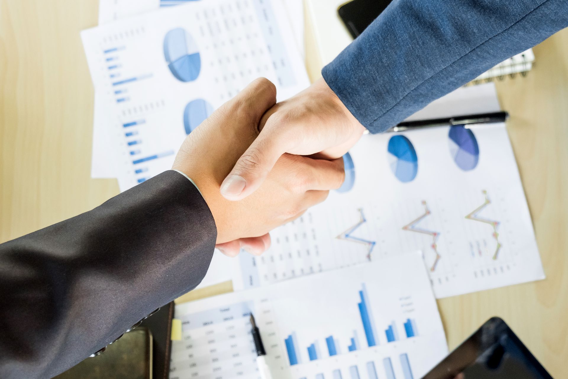 Two business people are shaking hands over a table with graphs on it.