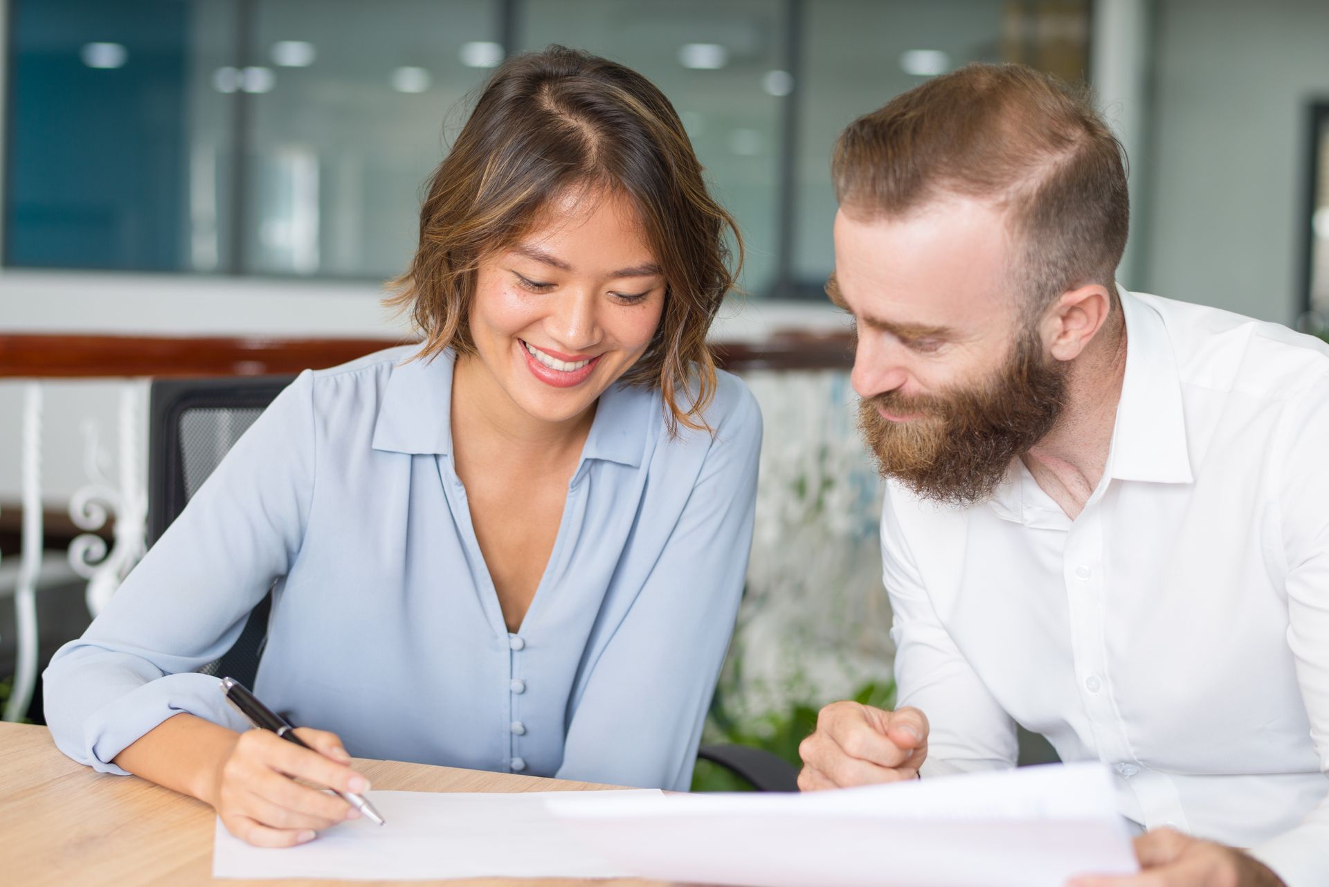 A woman and a man smiling as they review documents together in an office setting. The woman is writing.