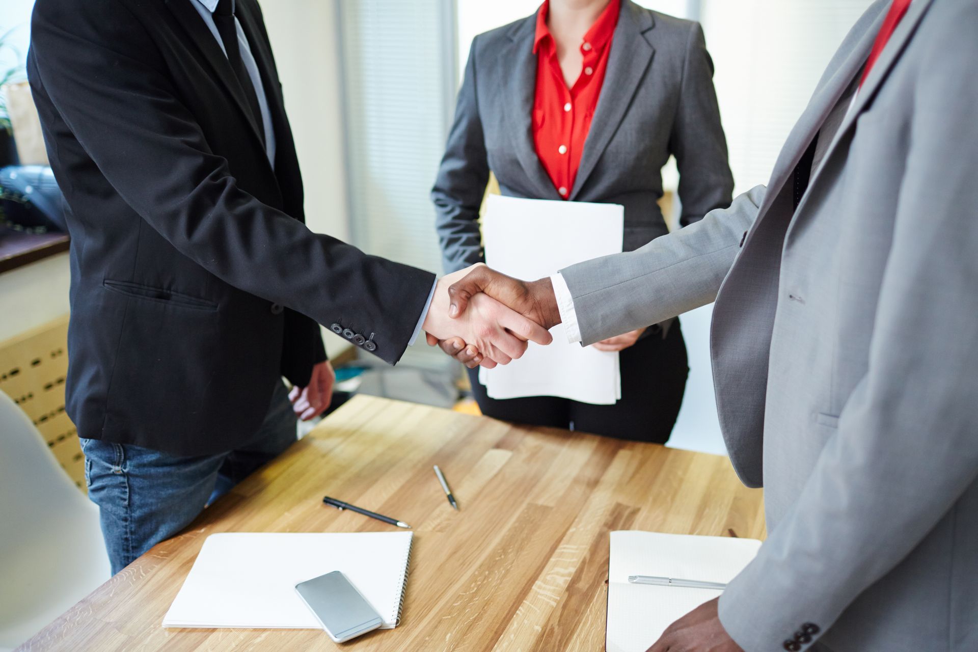 Two men shaking hands across a wooden table, a woman in a grey suit and red shirt stands behind them, holding papers.