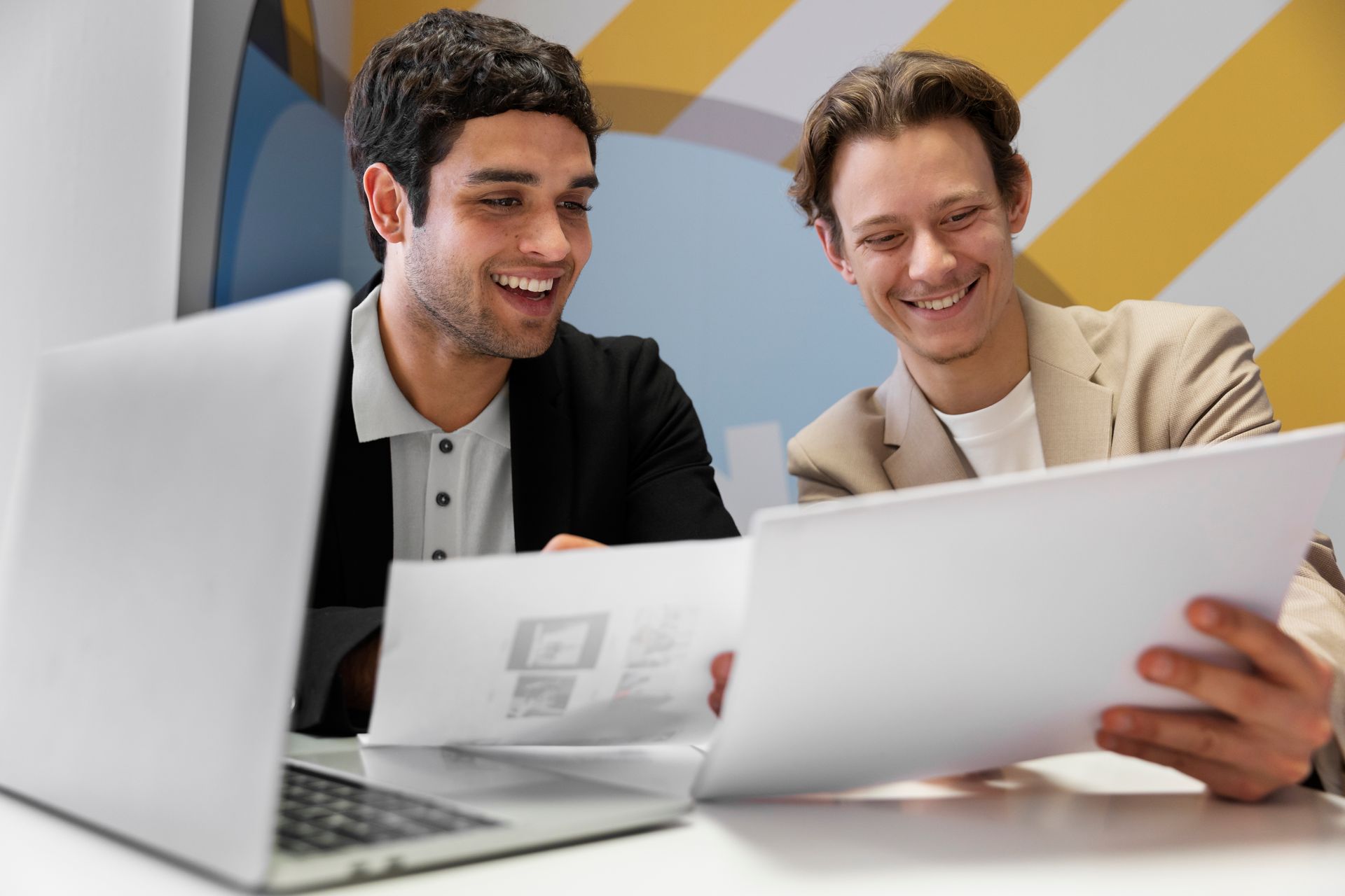 Two men in an office setting, smiling and looking at papers. One has a laptop open; they appear to be collaborating.