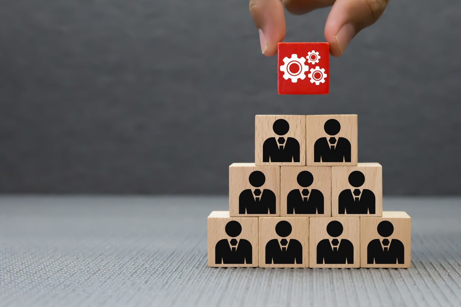 Hand placing a red block with gears on top of a pyramid of blocks with people icons, symbolizing teamwork.