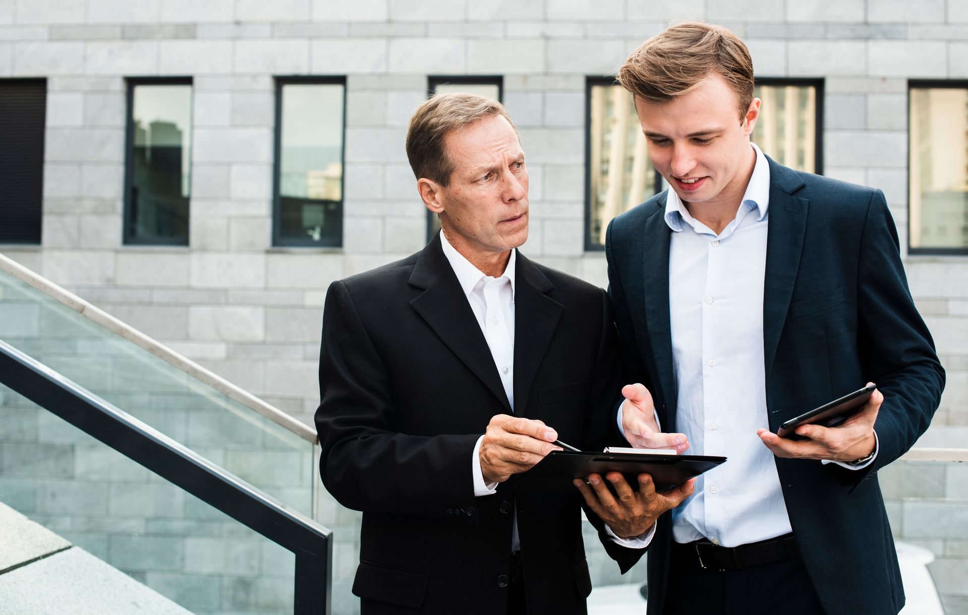 Two men in suits are standing next to each other looking at a tablet.