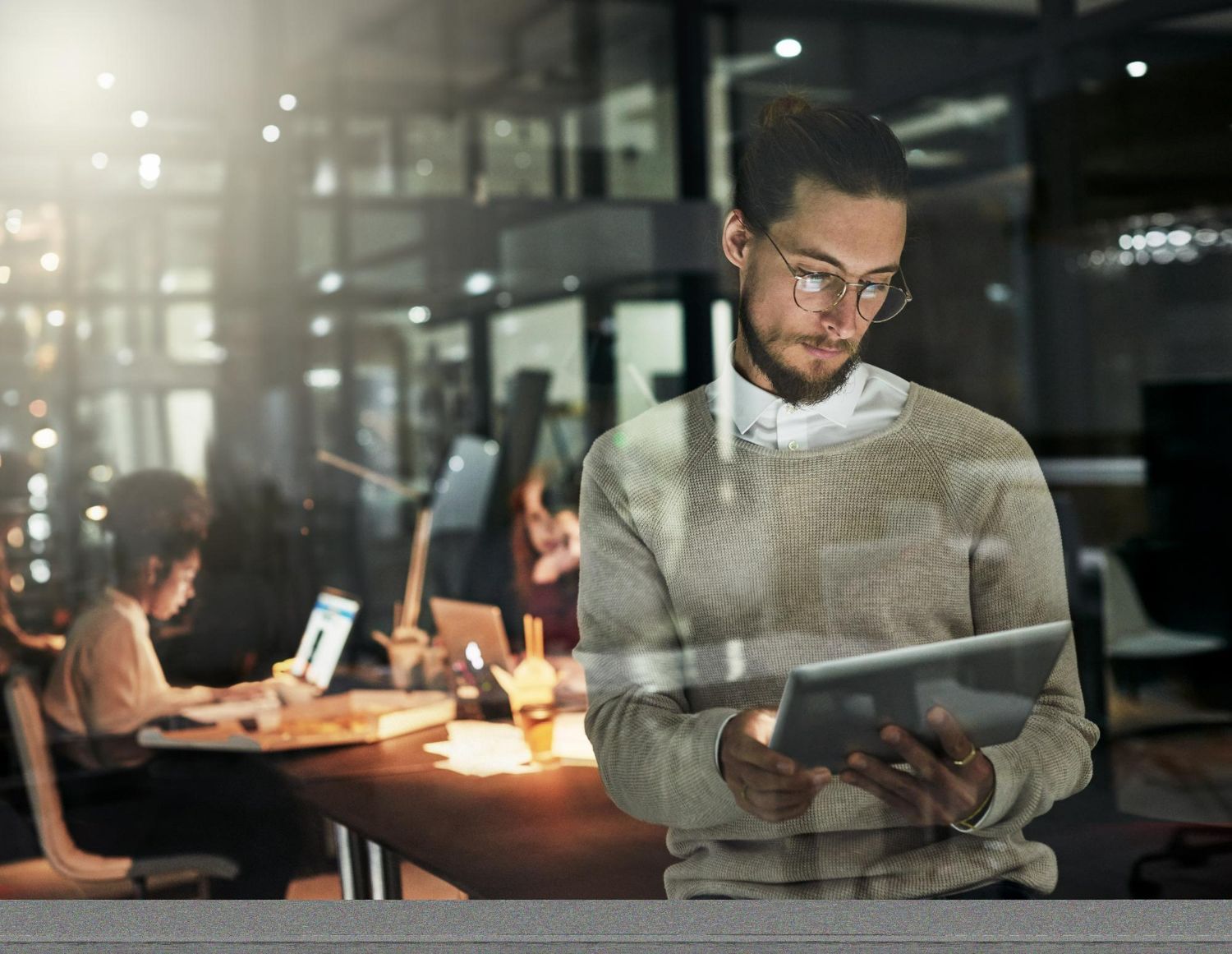 Man in glasses and sweater using a tablet in a dimly lit office. Another person works at a desk nearby.