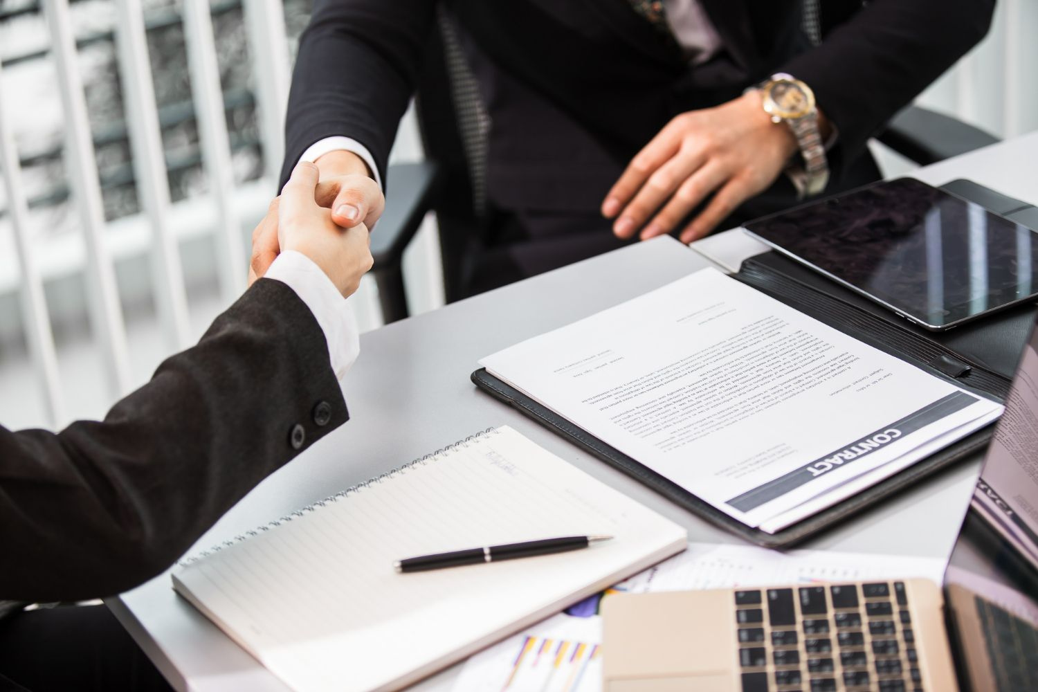 Two people in business suits shaking hands over a table with documents and a laptop.