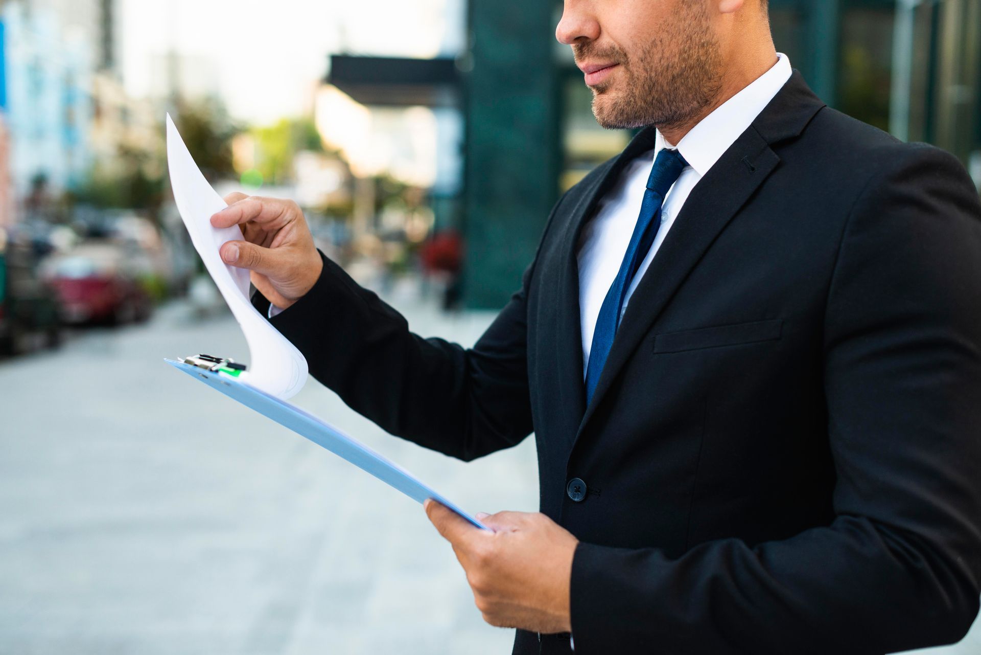 Man in suit examining papers on a clipboard outdoors. He's holding the top paper and looking down at it.