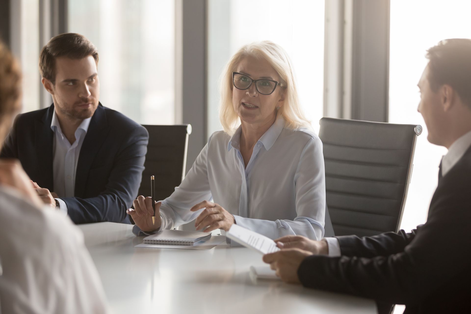 Business team in a conference room discussing documents. A woman with glasses gestures while speaking.