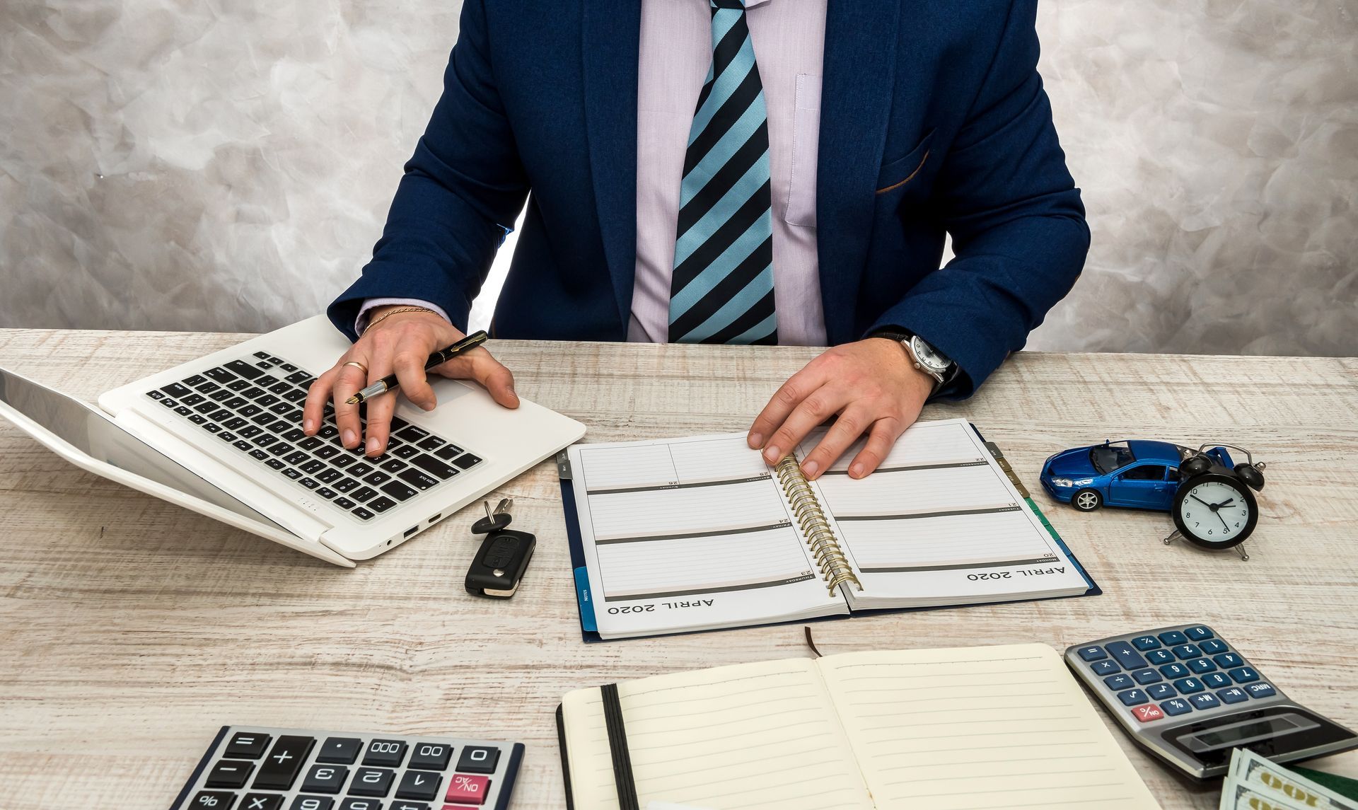 A person in a suit works at a desk with a laptop, planner, and calculator. A model car and clock are also present.