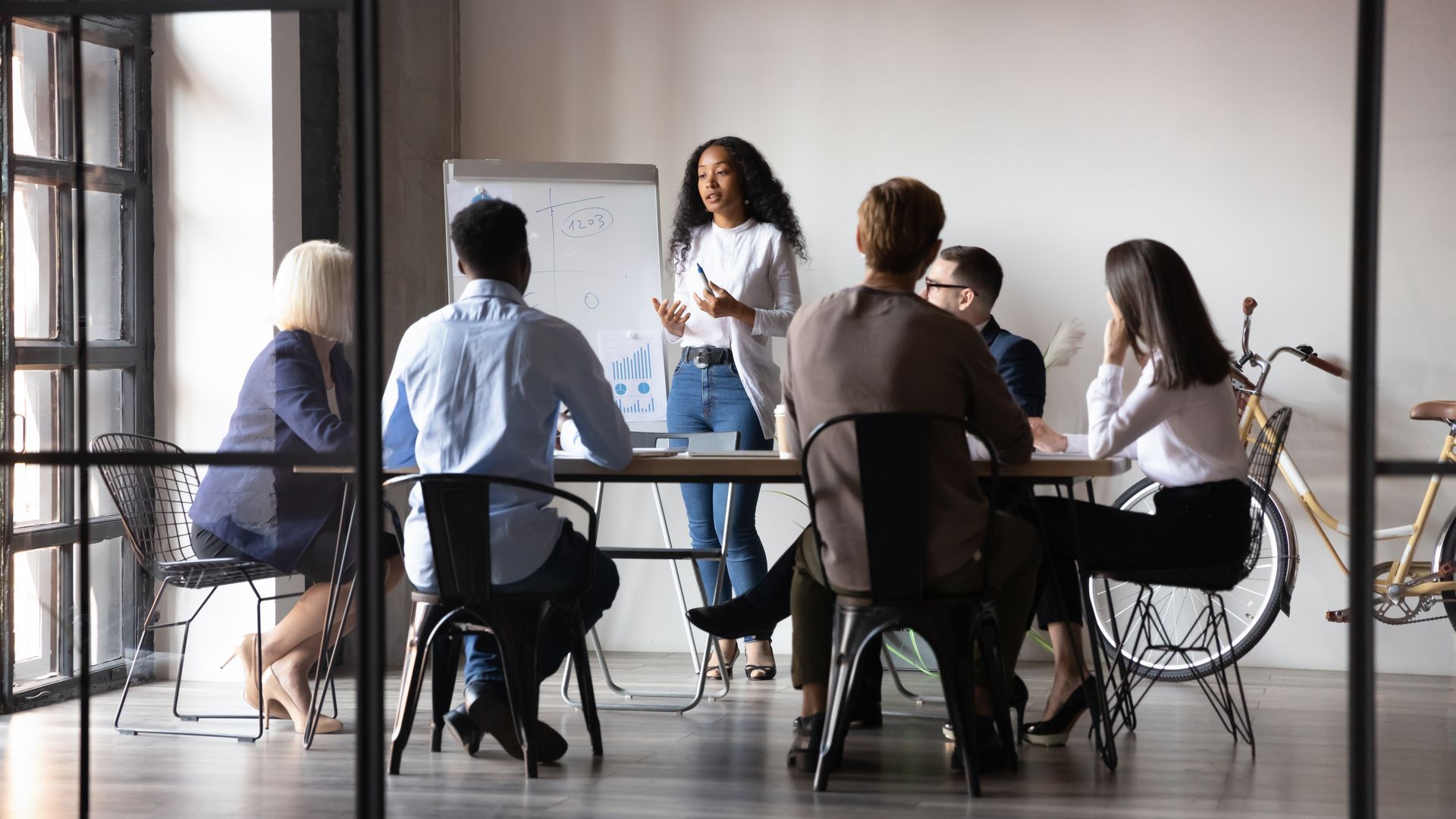 Group of diverse professionals in a meeting. Woman presents at whiteboard; others sit at table. Bright office setting.