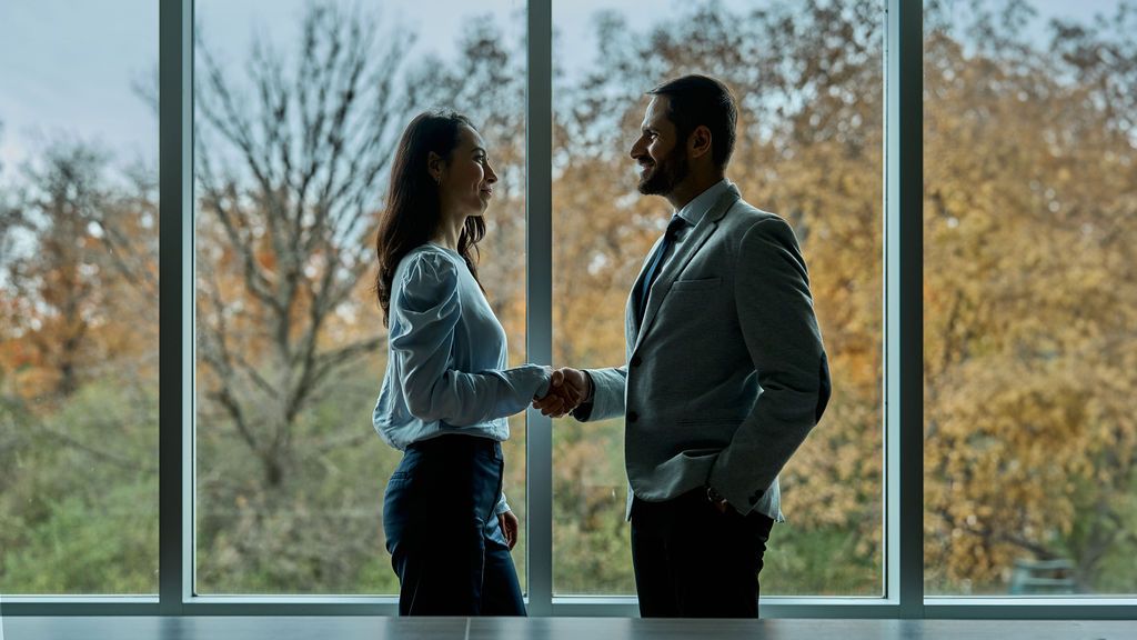 A man and a woman are shaking hands in front of a window.