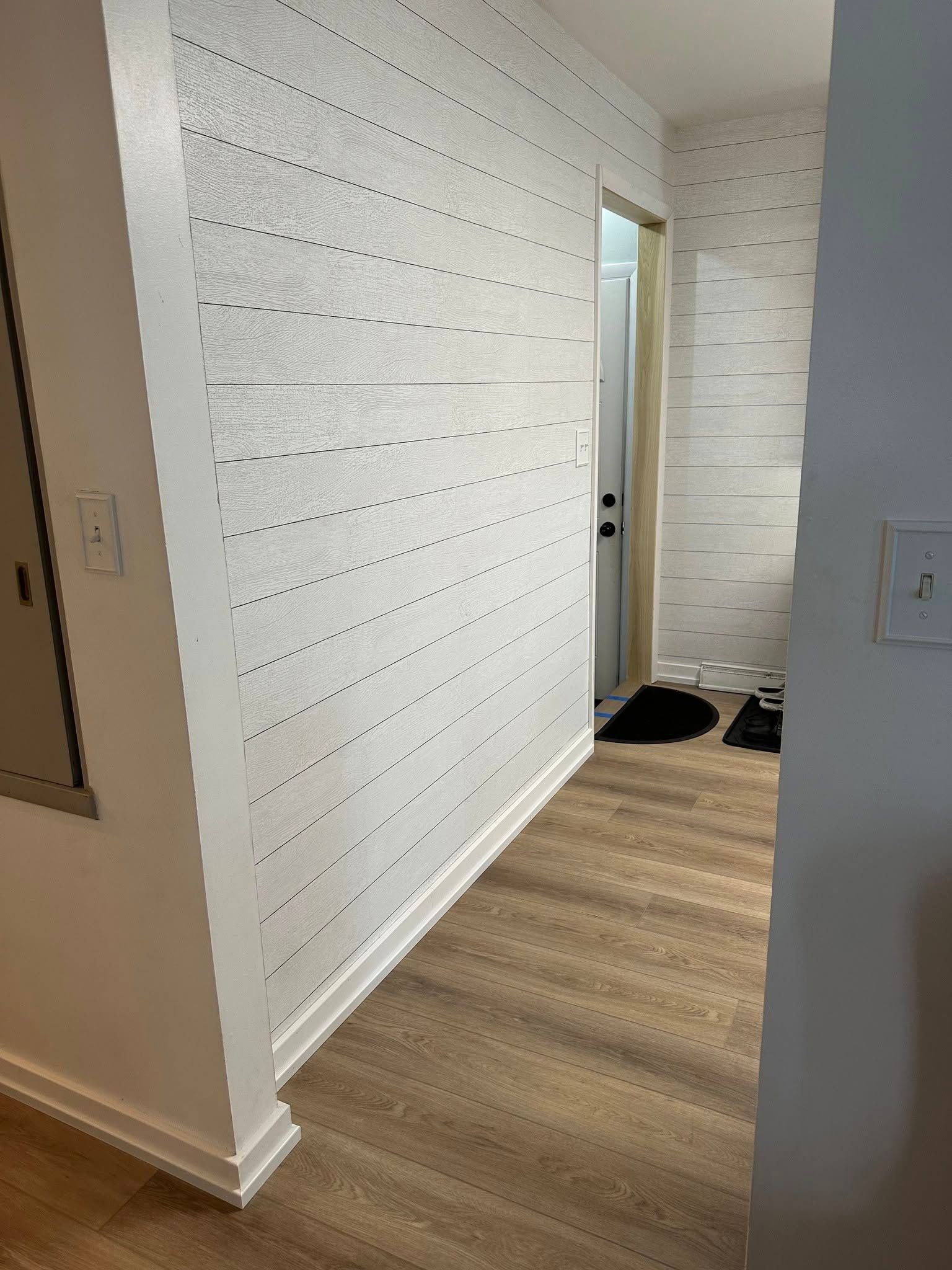 A hallway with a white wall and wooden floor leading to a bathroom.