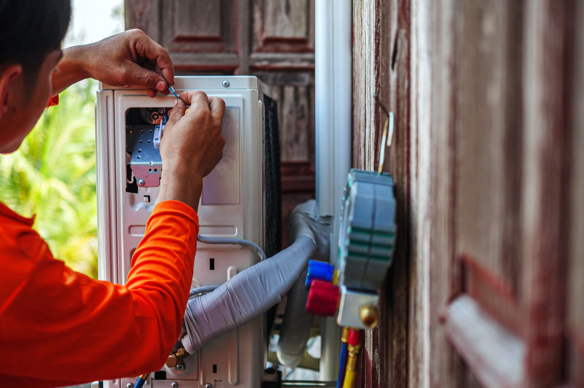 Technician installing an AC unit on a wooden wall, hands working inside, orange shirt.