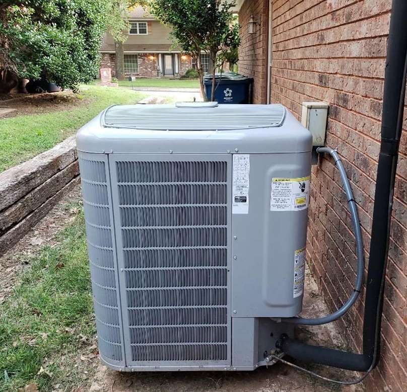 An air conditioner is sitting on the side of a brick building. An air conditioner is sitting in the grass in front of a brick wall.