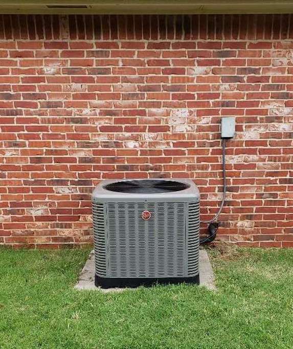 An air conditioner is sitting in the grass in front of a brick wall.