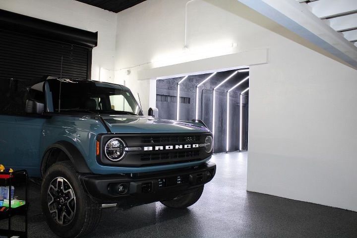 A teal Ford Bronco parked inside a well-lit garage; focus on the front, with a unique lighting display in the background.