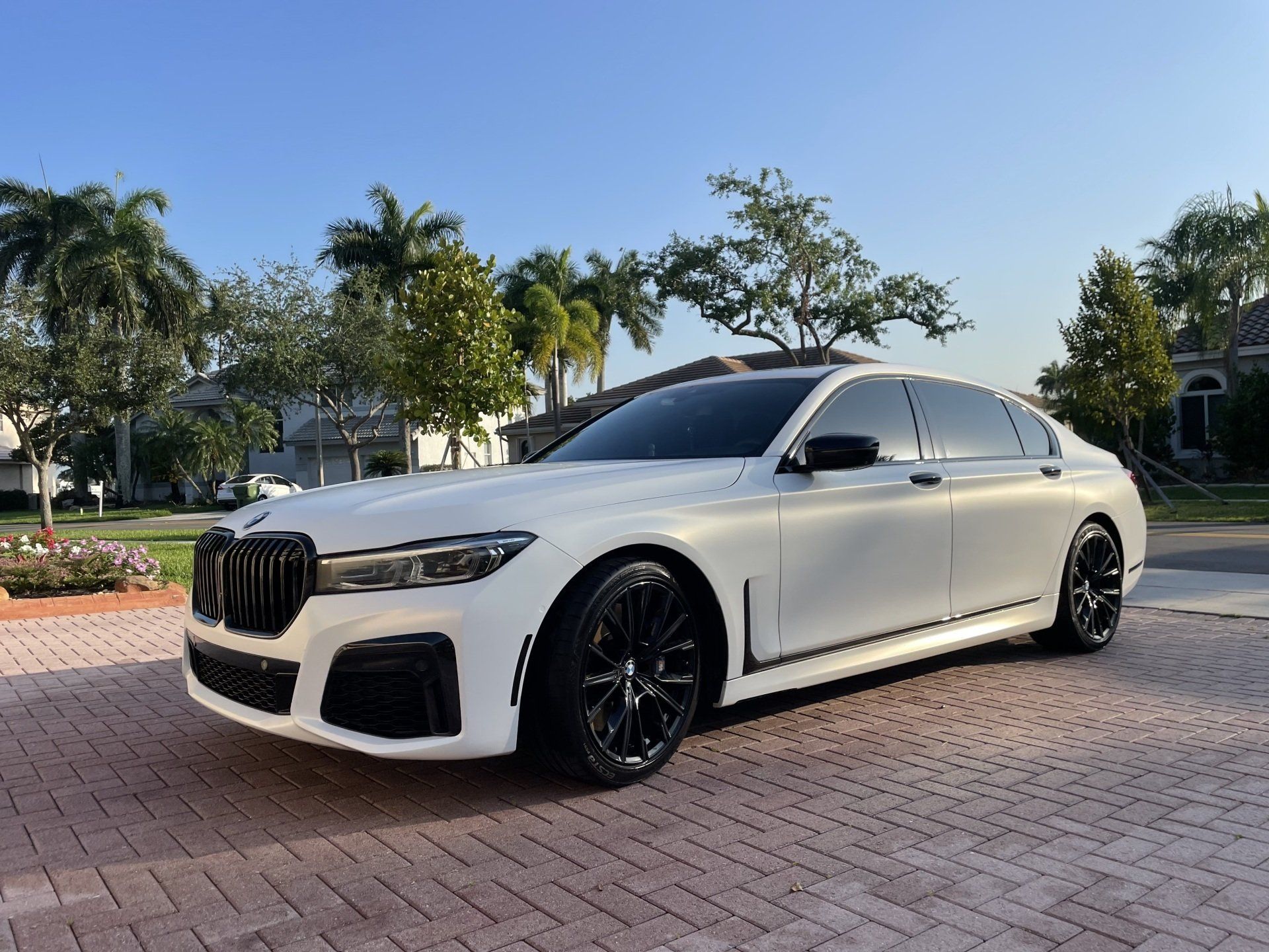 White BMW 7 series sedan parked on a brick driveway, black wheels, sunny day, trees and houses in background.