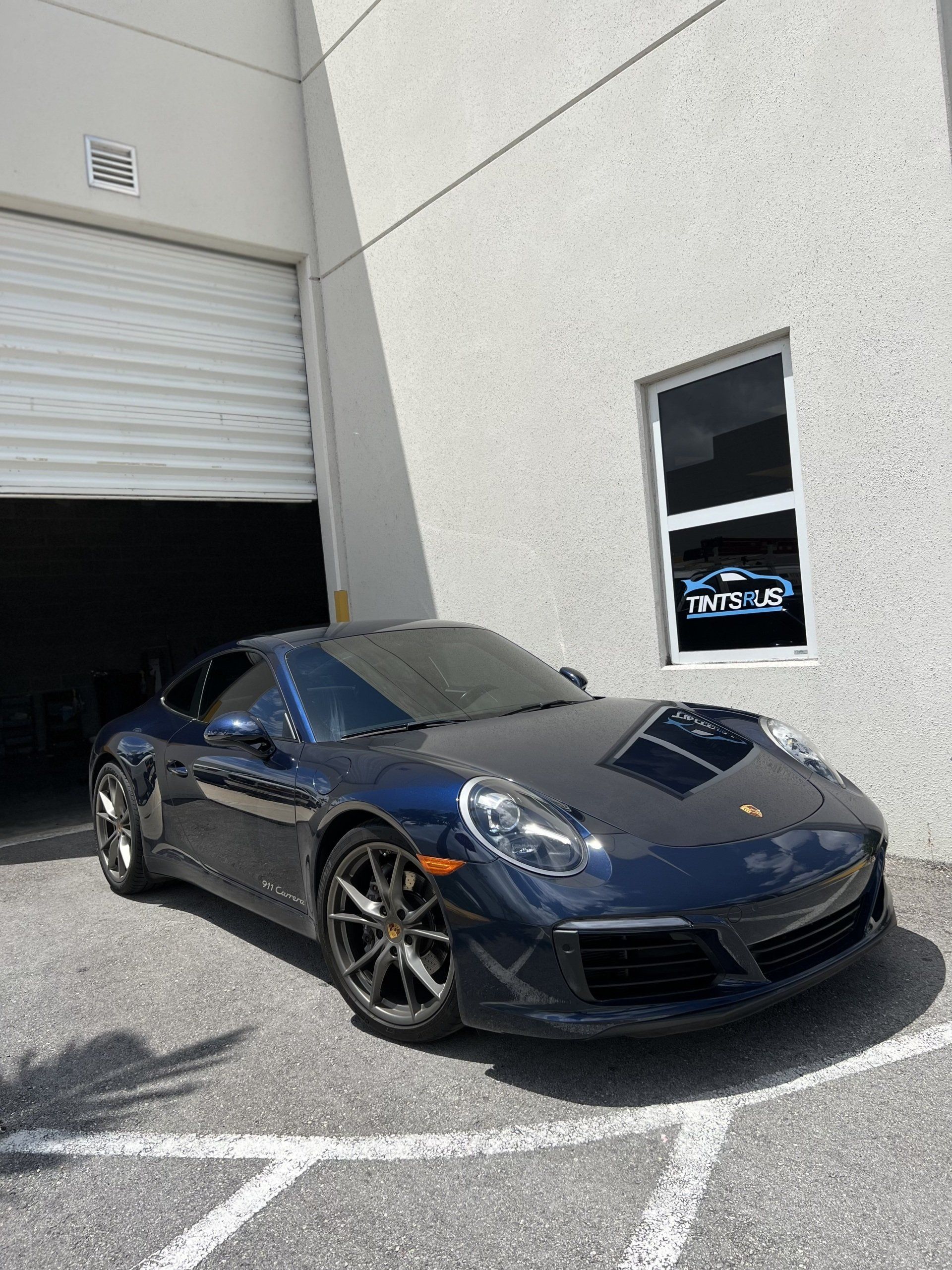Dark blue Porsche parked outside a white building with a closed garage door, sunny day.