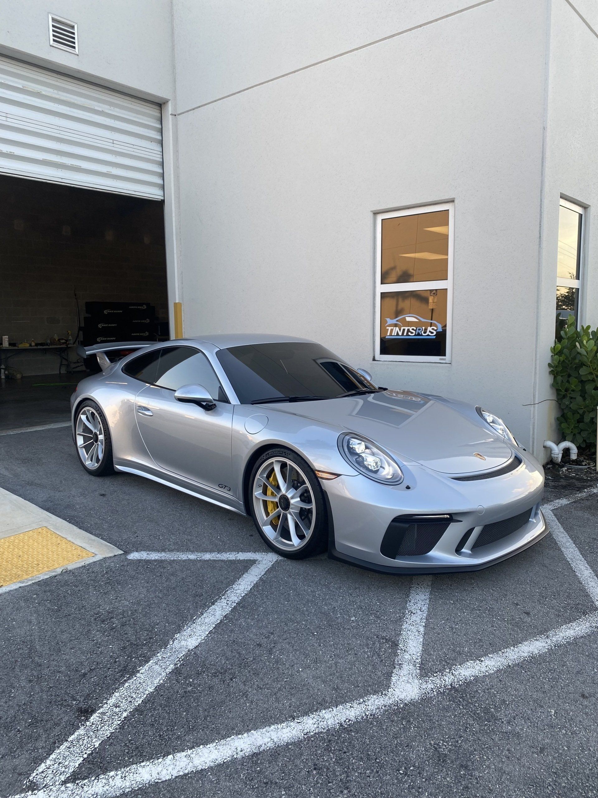 Silver Porsche 911 parked outside a light gray building with a garage door, in daylight.