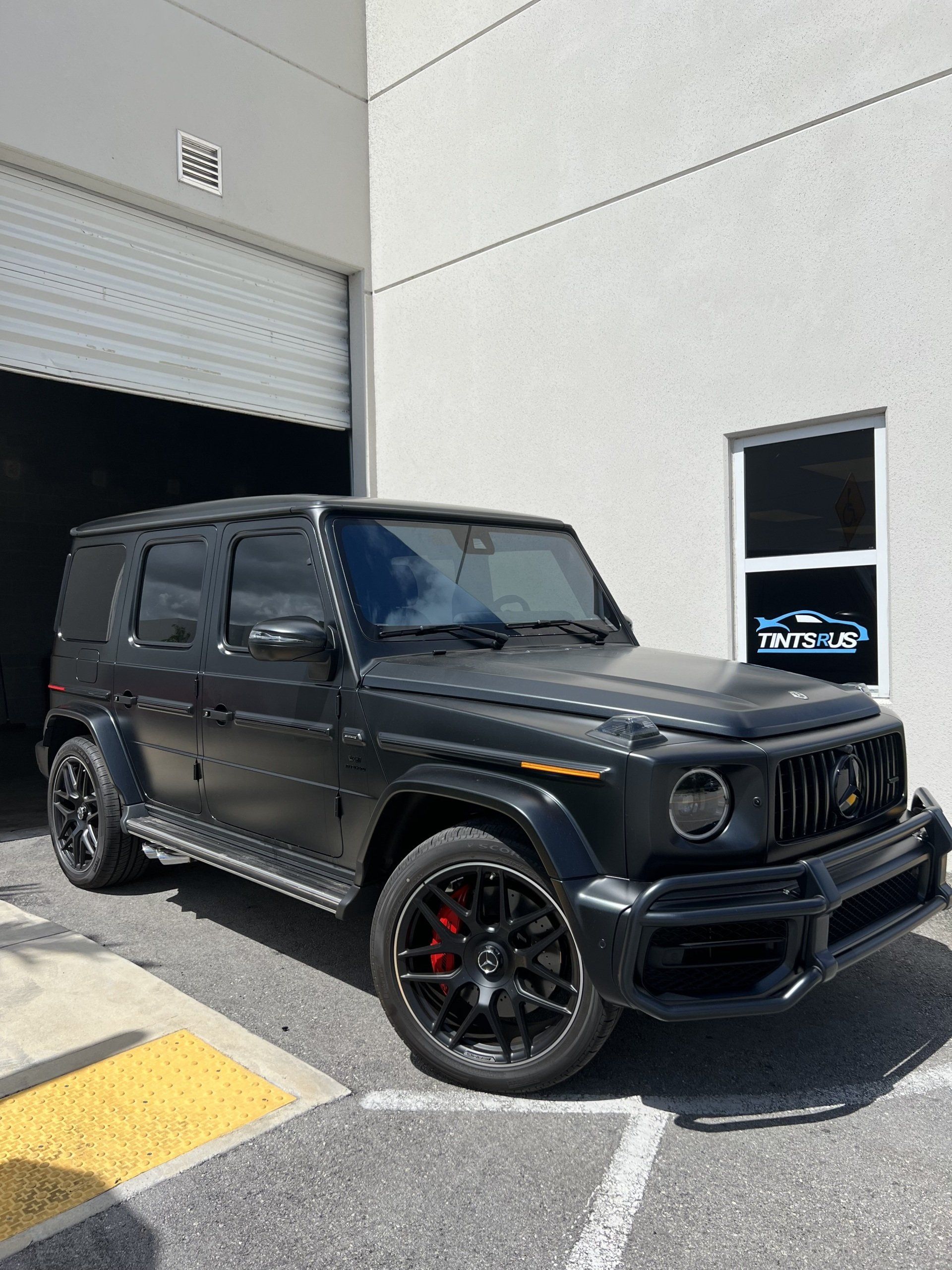 Black Mercedes-Benz G-Wagon parked outside a building with a roll-up door and a window displaying a logo.