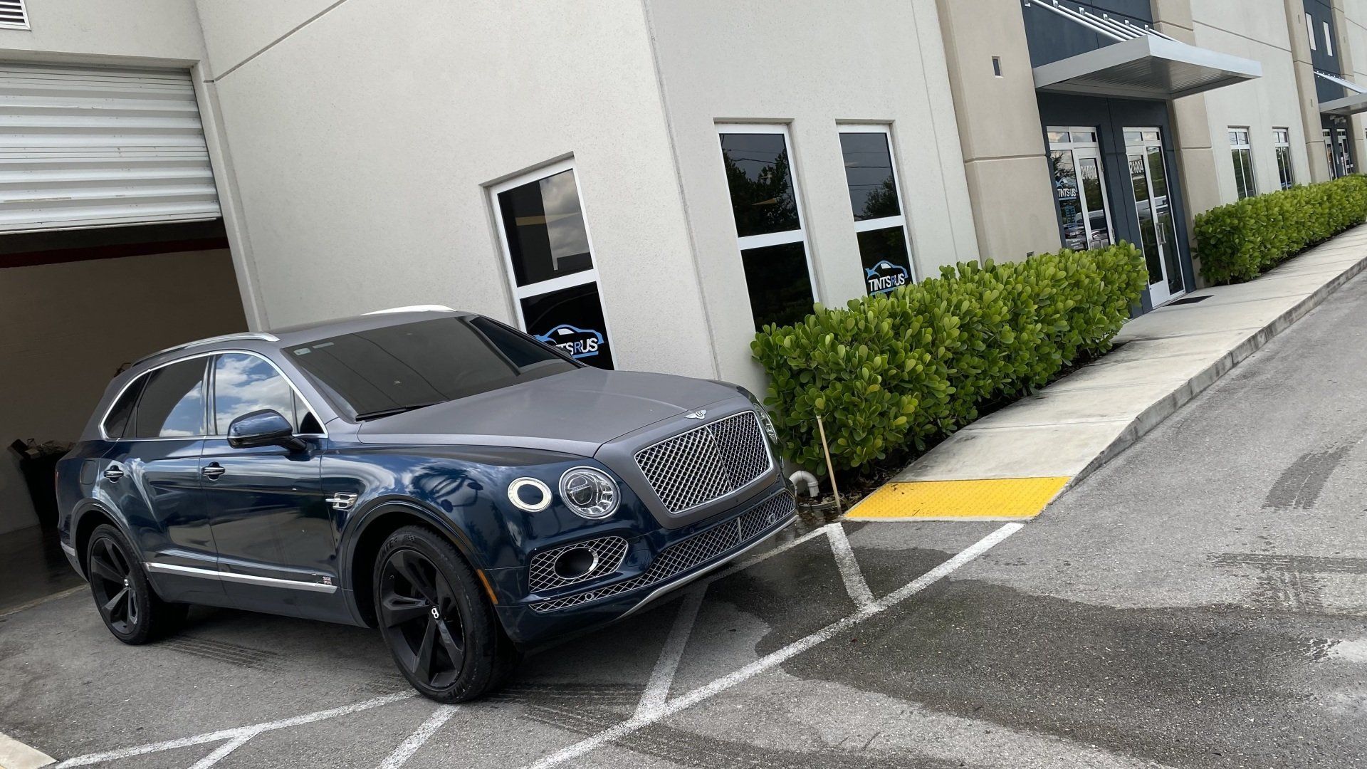 Dark blue Bentley SUV parked in front of a modern building with bushes and a yellow curb.