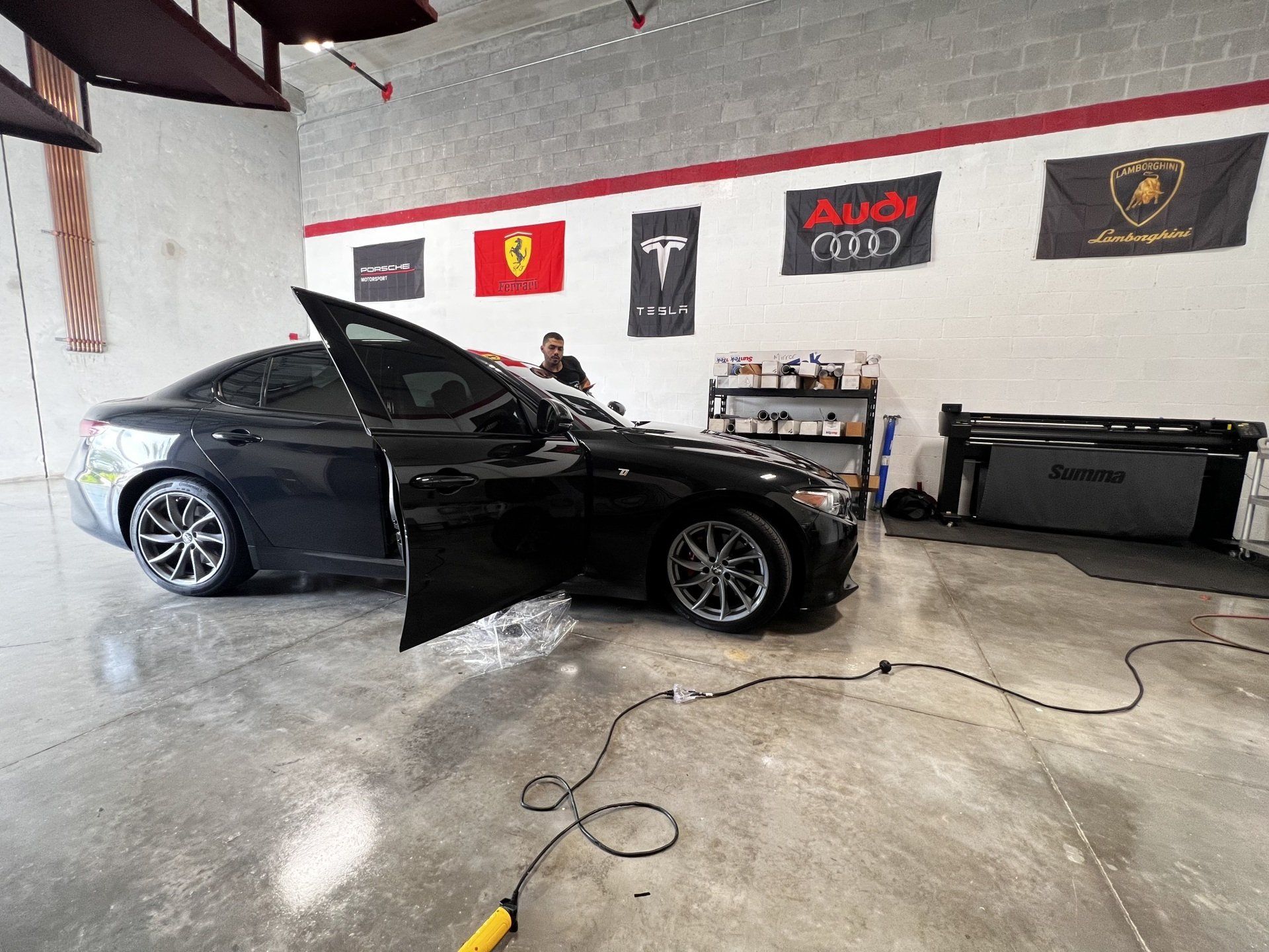 Black car with open door in a shop, being worked on. Various car brand flags displayed.