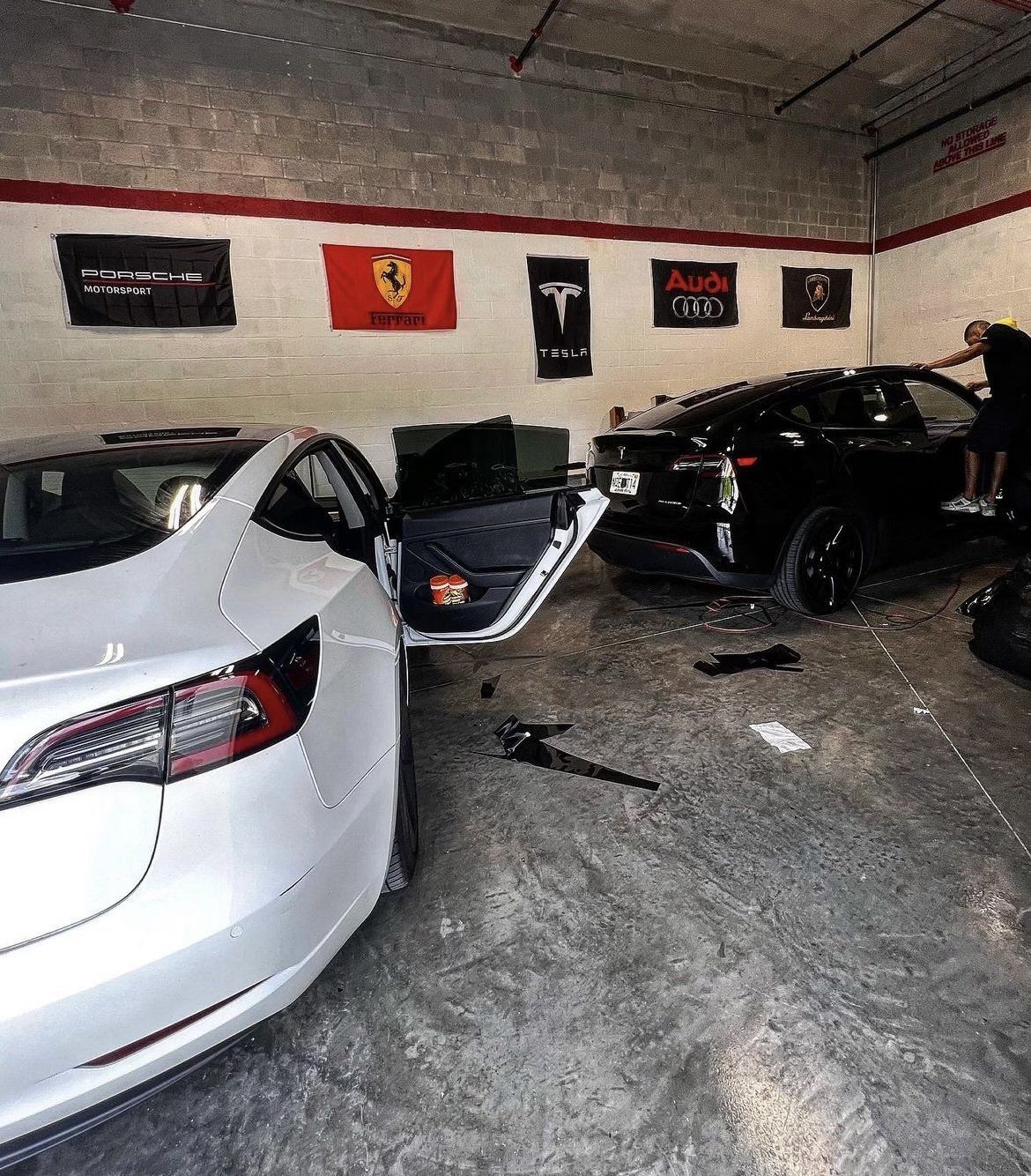 A white Tesla and black car in a garage. A person works on the black car. Flags hang on the wall.