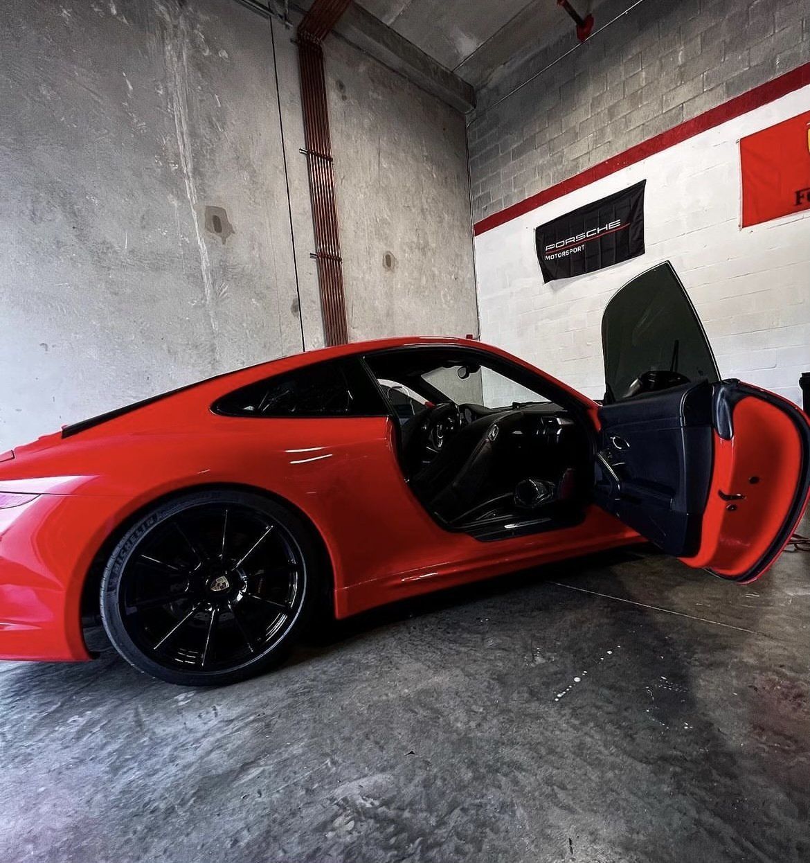 Red Porsche 911 sports car with black wheels and open door parked inside a garage.