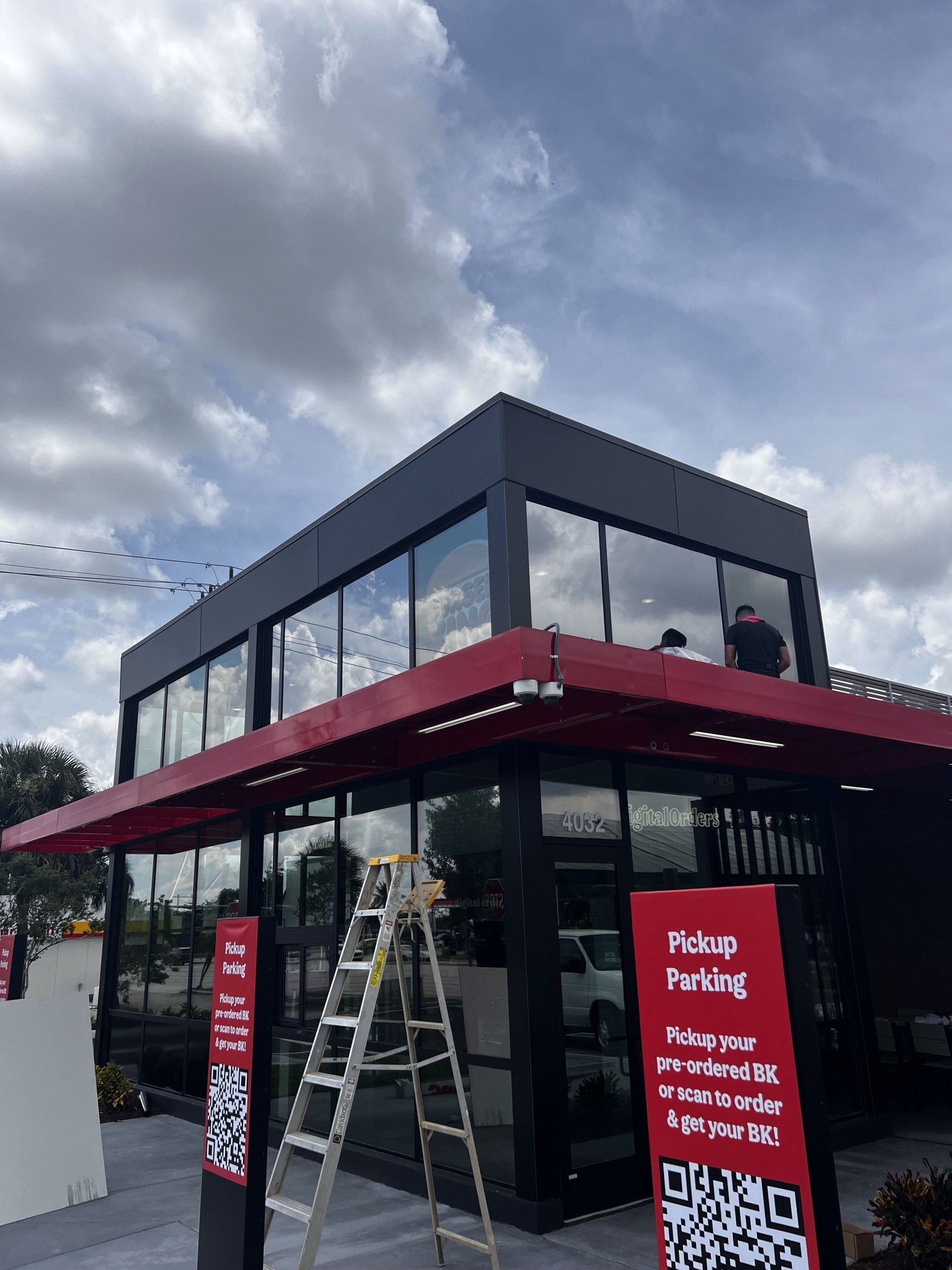 Modern restaurant exterior with red accents, cloudy sky. Pickup parking sign, ladder.