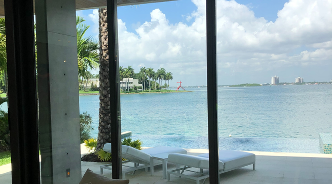 Exterior view of a modern home with a pool overlooking a bay, white lounge chairs, blue water, and a sunny sky.