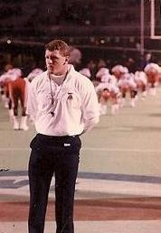 A man is standing on a football field in front of a row of football players.