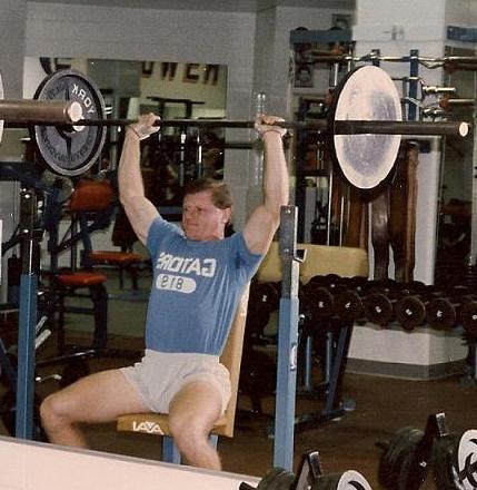 A man lifting a barbell in a gym wearing a shirt that says rotaj