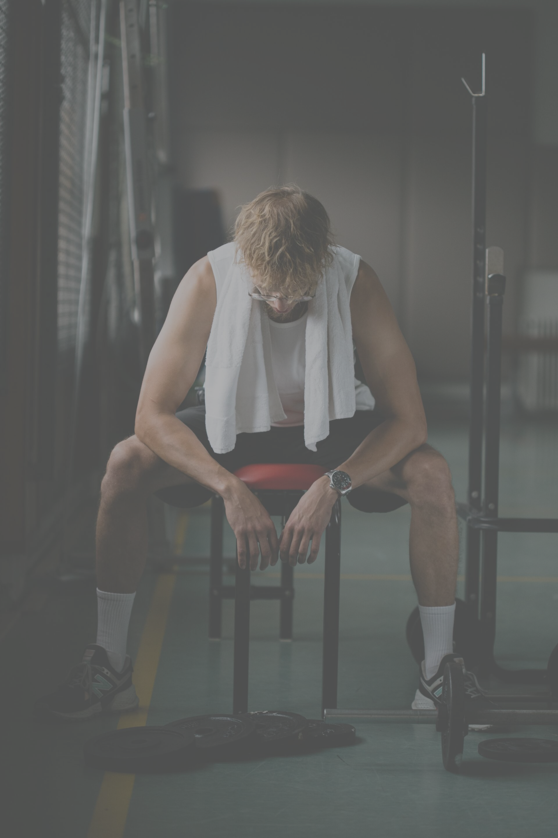 A man is sitting on a bench in a gym with a towel around his neck.
