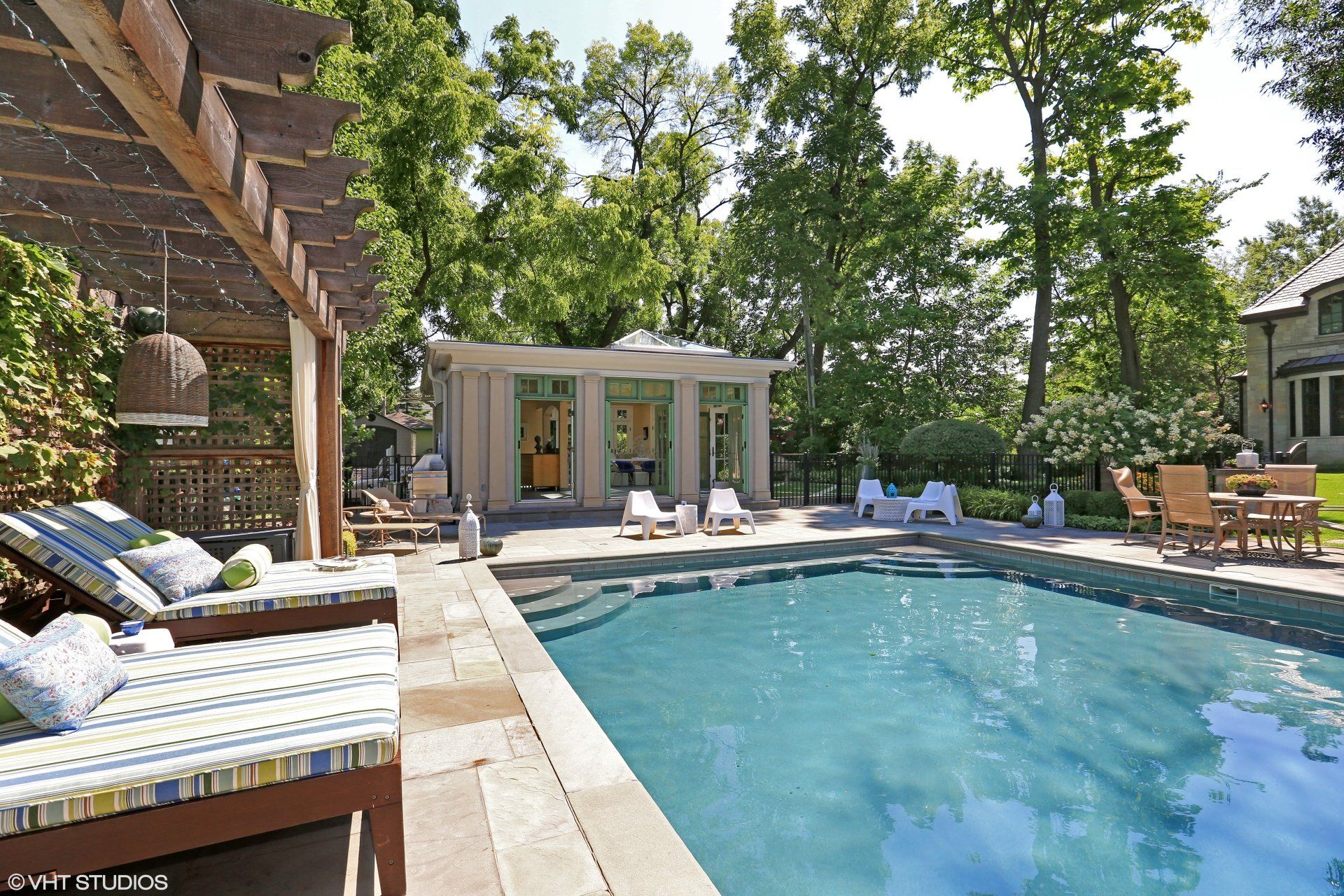 A large swimming pool is surrounded by chairs and a pergola