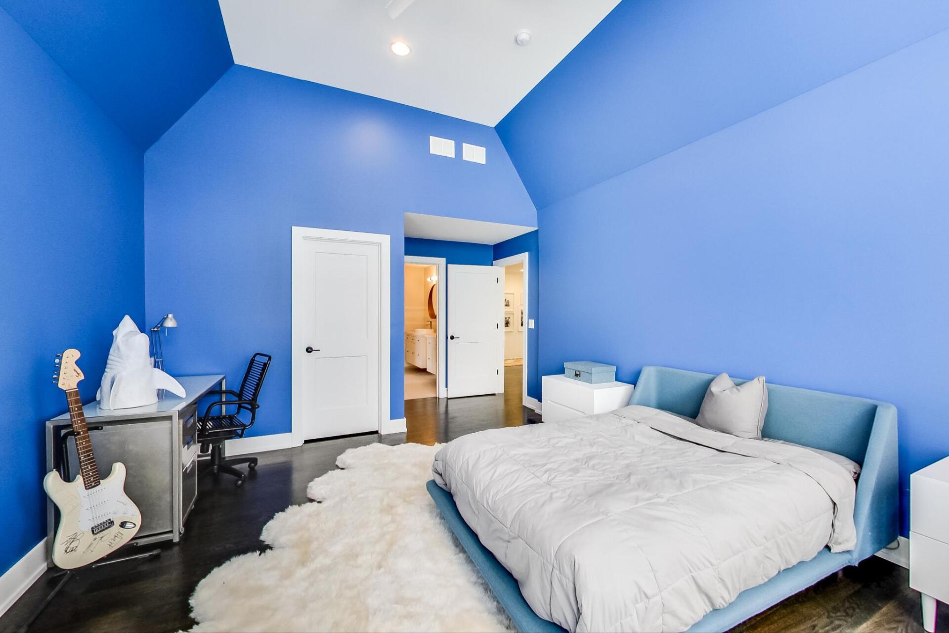 A bedroom with blue walls , a bed , a desk and a guitar.