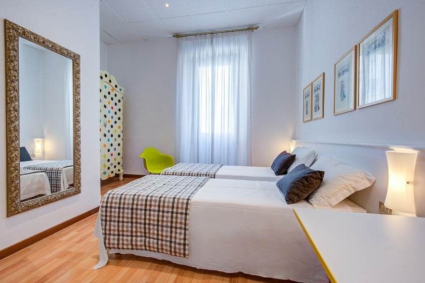 Un dormitorio luminoso con dos camas, un gran espejo, un biombo y una ventana con cortinas transparentes.