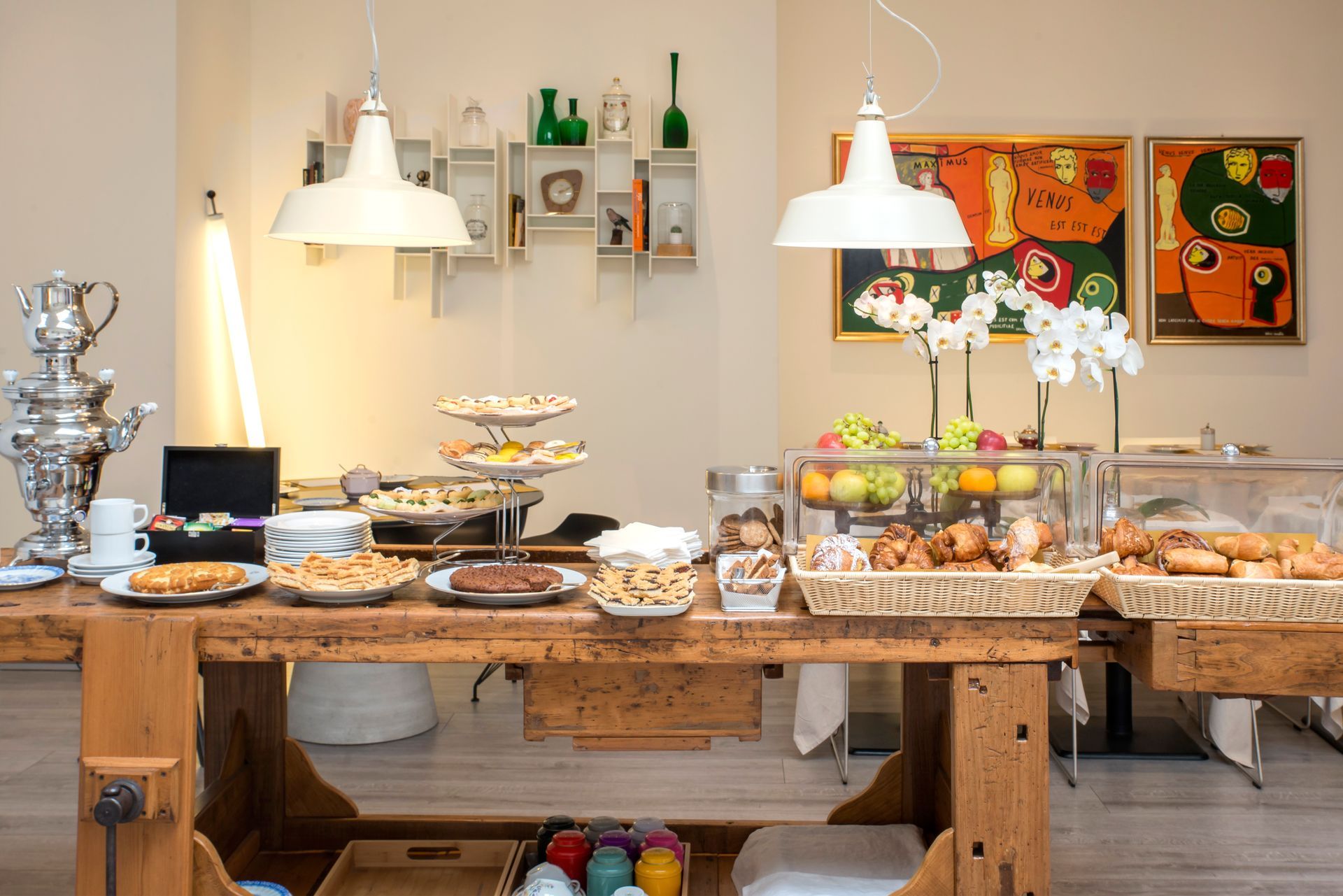 Breakfast buffet: pastries, fruit, and snacks on a wooden table, under pendant lights, with art on the wall.